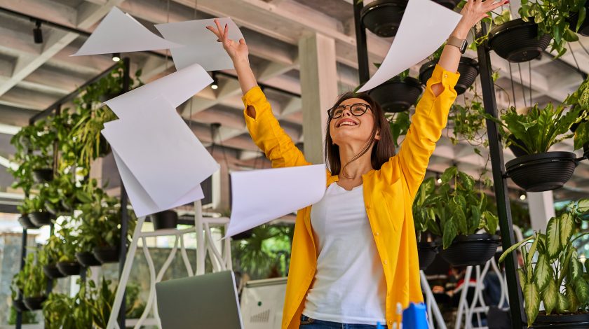 A woman joyfully throws papers into the air inside a modern, green office space adorned with lush hanging plants and natural light.