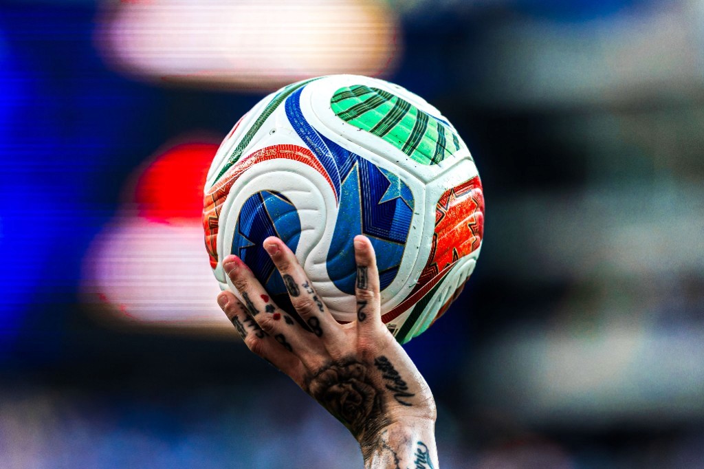 A tattooed hand holds the World Cup match ball aloft in the stadium lights