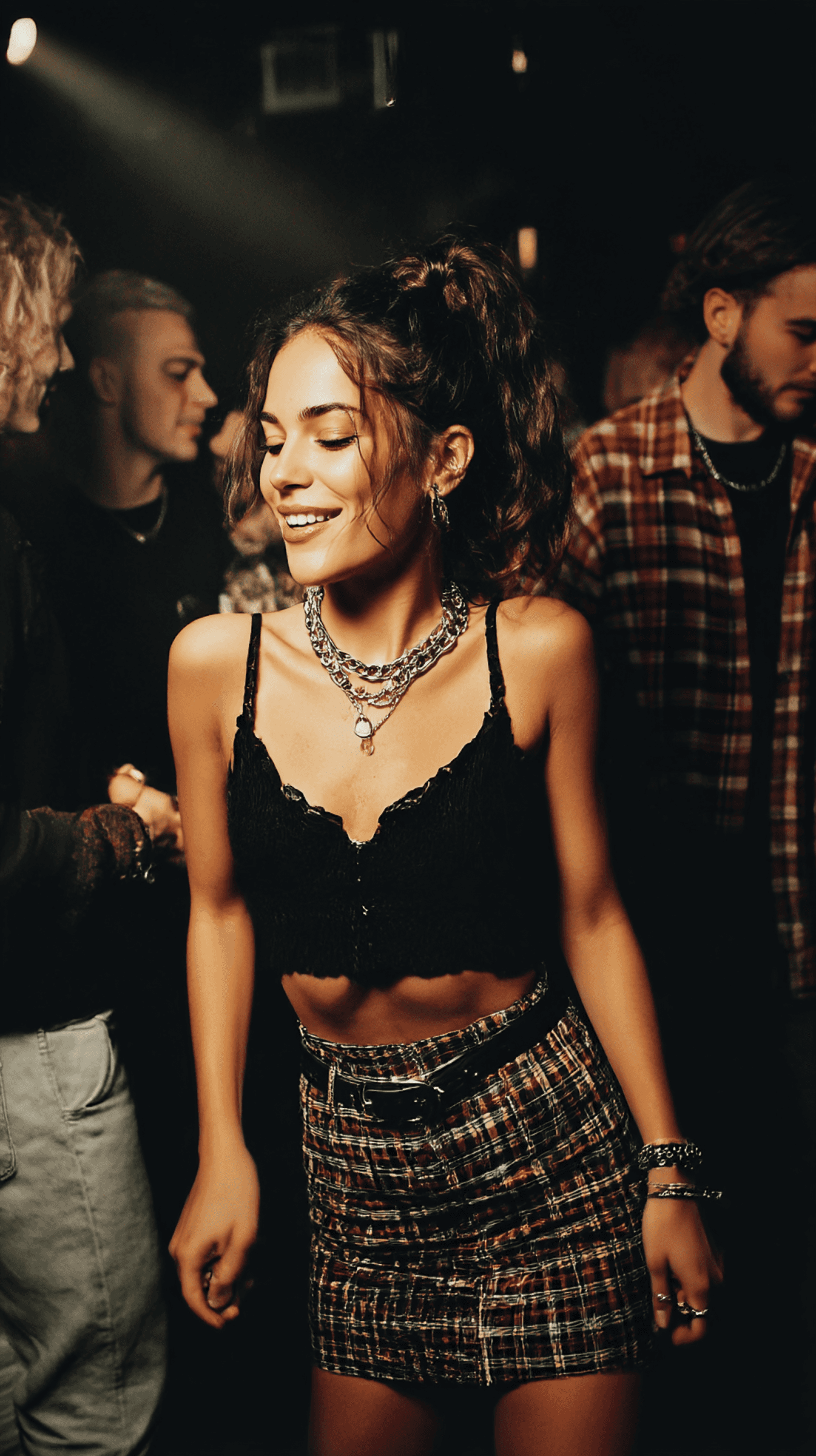 A young woman in a black lace top and plaid skirt enjoys a lively party atmosphere, surrounded by friends in a dimly lit setting.