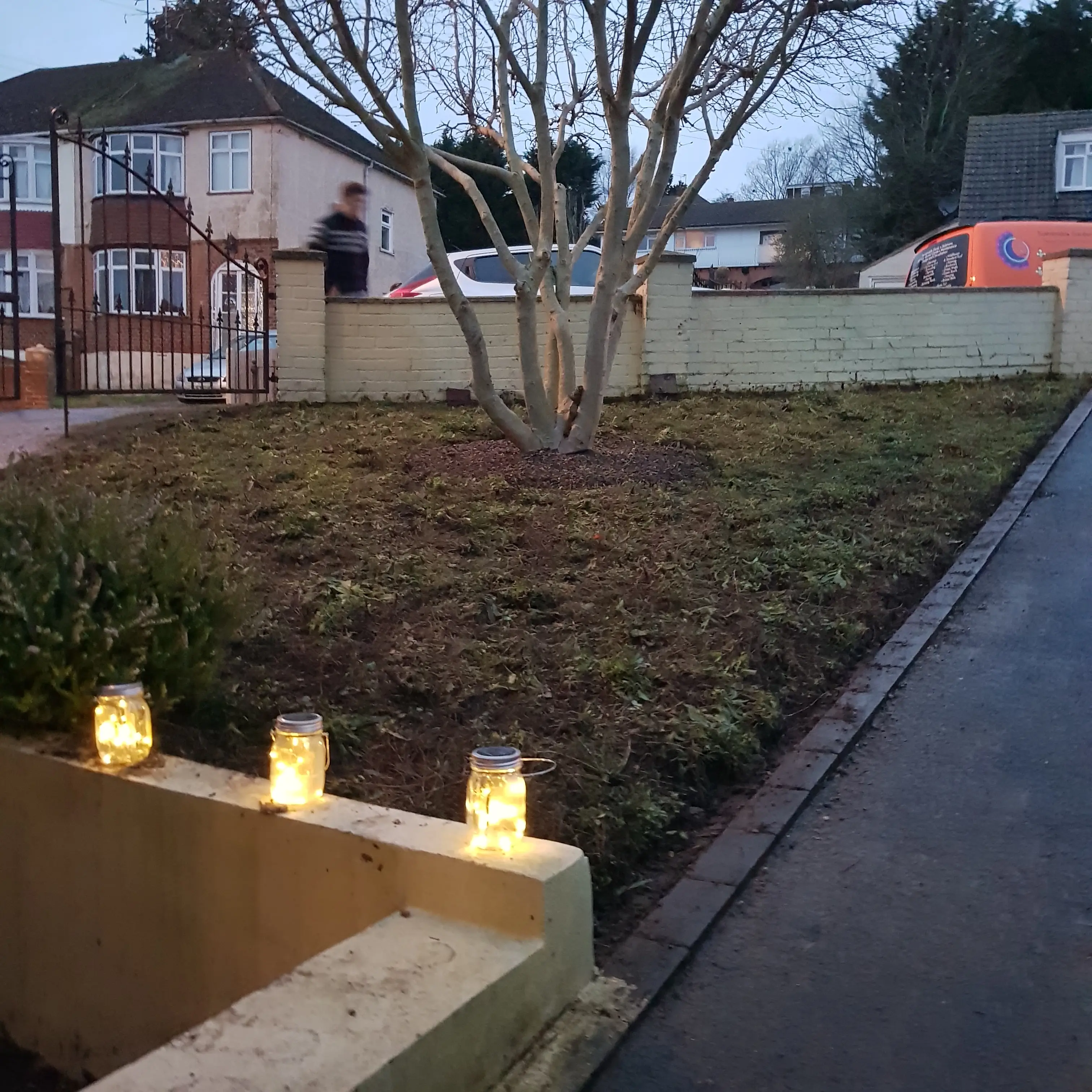 A residential garden with a sloped walkway, a small tree, and lanterns along the path in soft evening light.