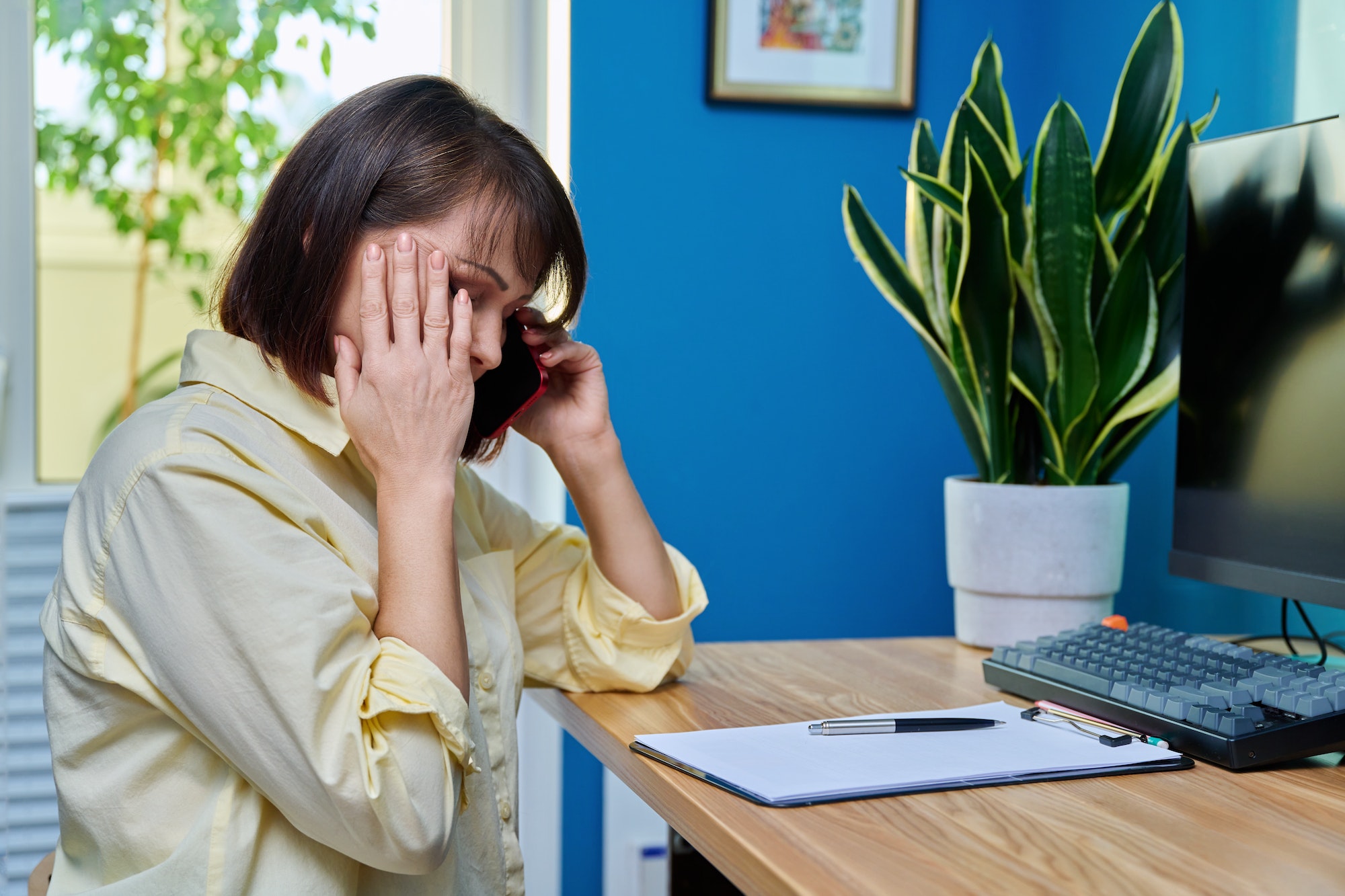 A woman at a desk looking distressed while talking on a red smartphone, with her hand covering her forehead