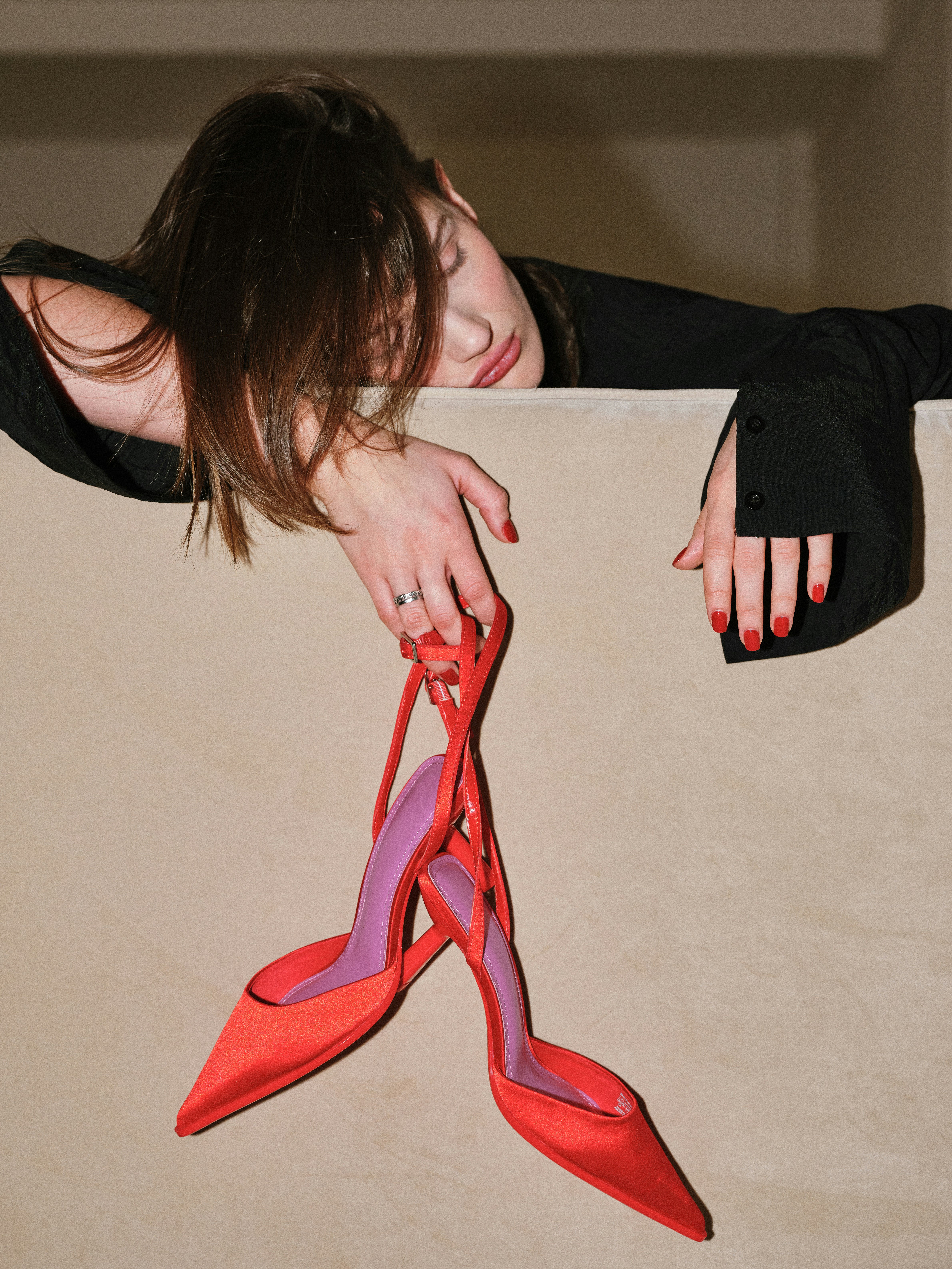 Languid editorial shot of a brunette model in a black shirt draped over a cream surface, dangling red satin strappy heels with purple insole from one hand
