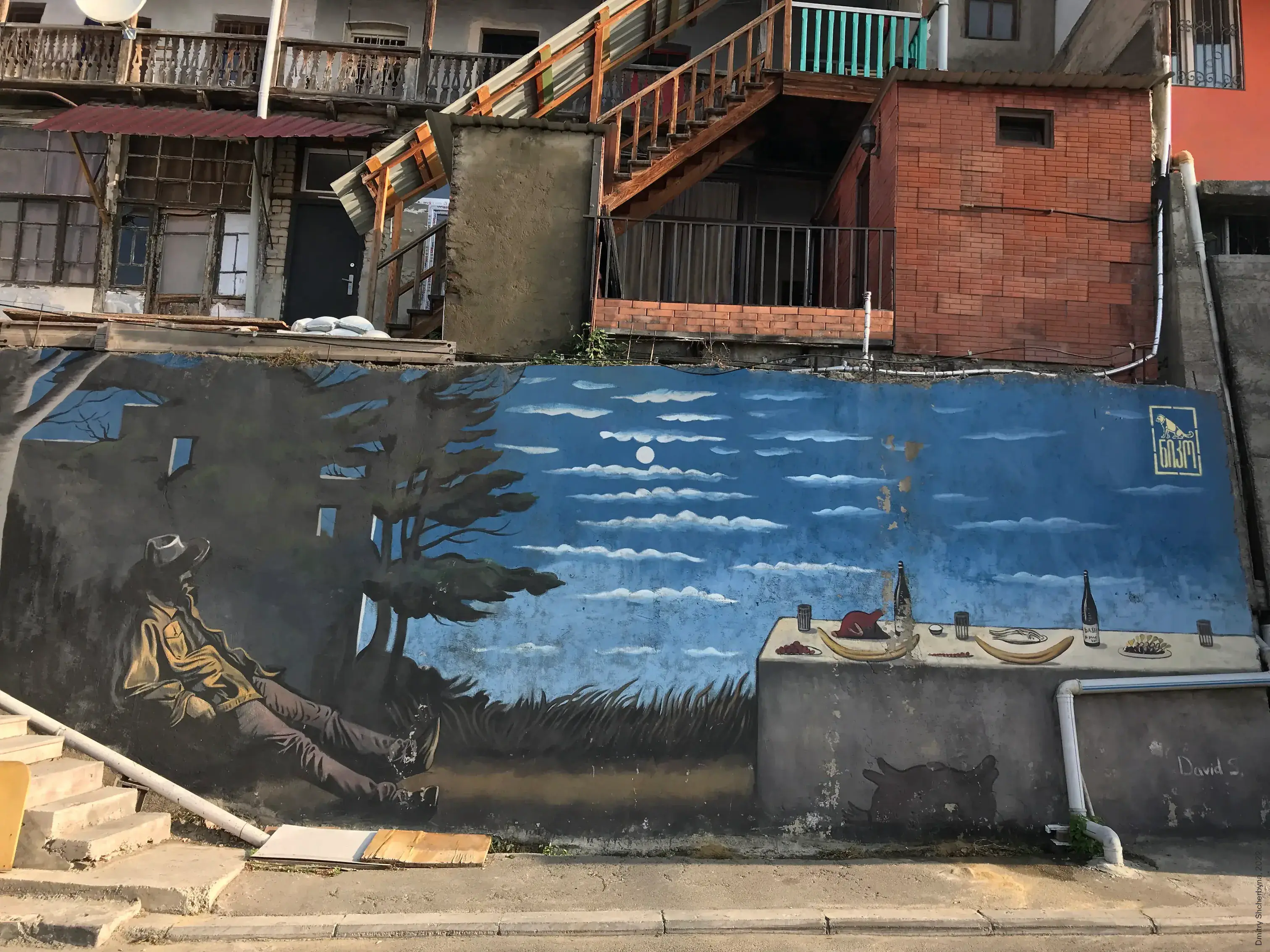 A mural on a residential wall shows a figure in a hat reclining beneath dark trees on the left, with a table set with wine bottles, food, and dishes on the right under a blue moonlit sky.