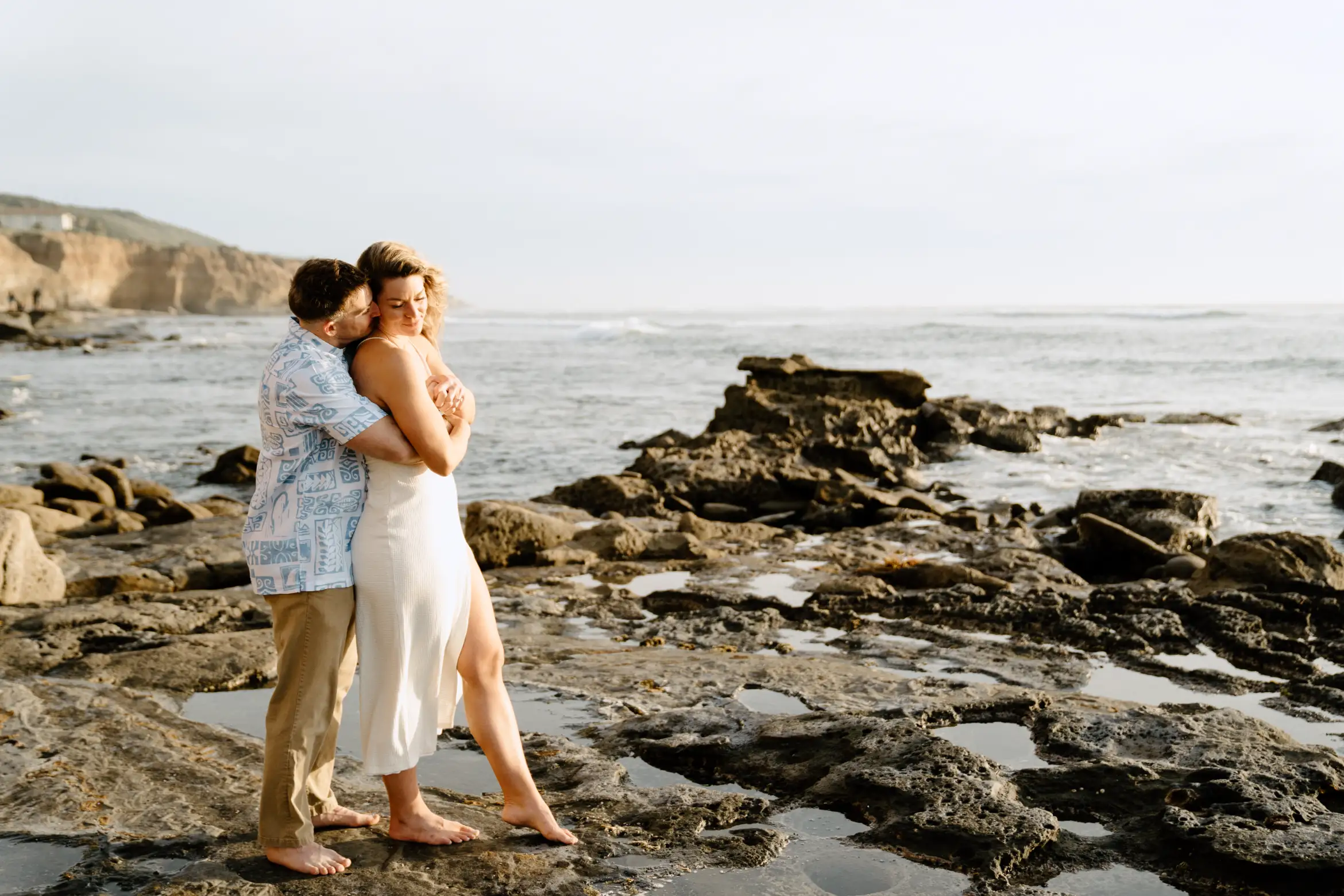 Two individuals embrace on a rocky beach, with calm waters and a hazy sky in the background.