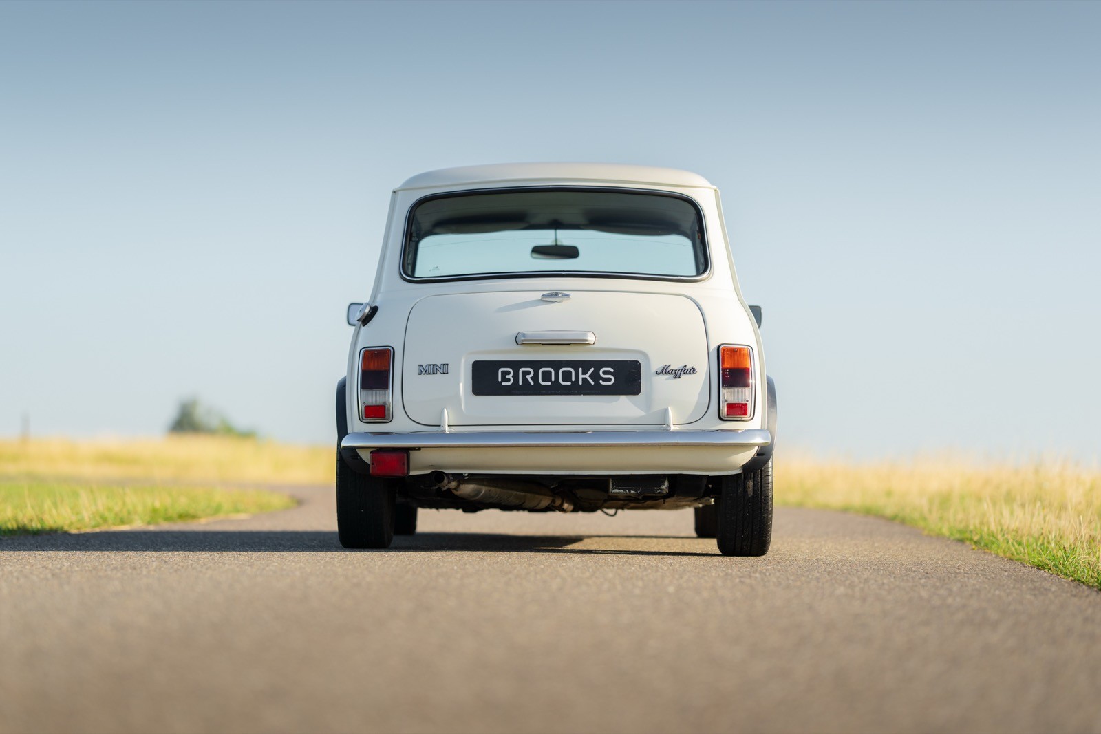 A rear view of a vintage car driving on a rural road, with open fields and a clear sky in the background.