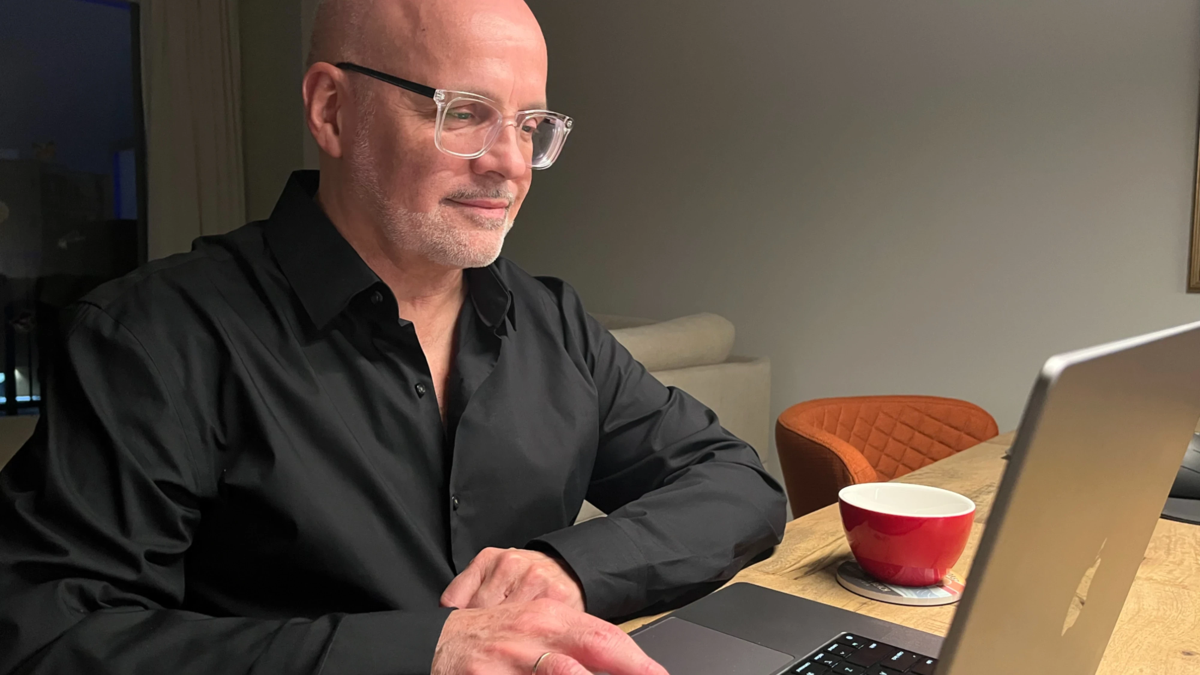 Stéphane Paquet working on a laptop at a wooden table with a red coffee cup beside him in a warmly lit room.