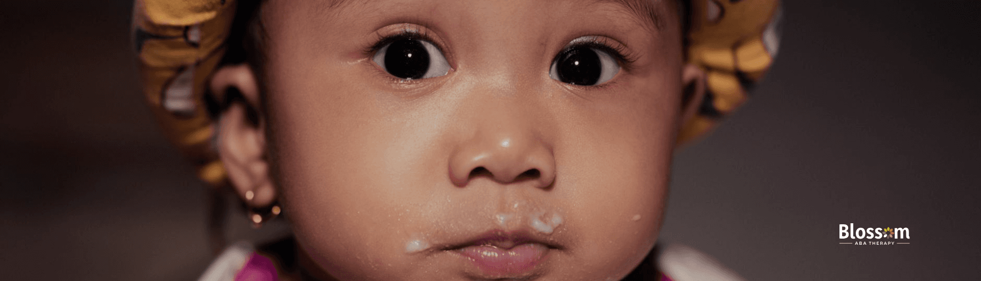 A toddler with autism and large eyes wearing a patterned headband, with food around their mouth.