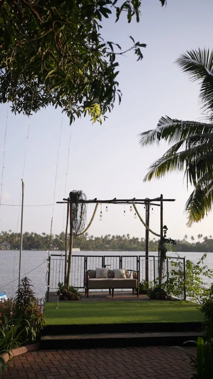 Riverside ceremony deck at Bhūmi courtyard, Vedic Village Kodungallur, with a seating swing, string lights and coconut palms by the Periyar River