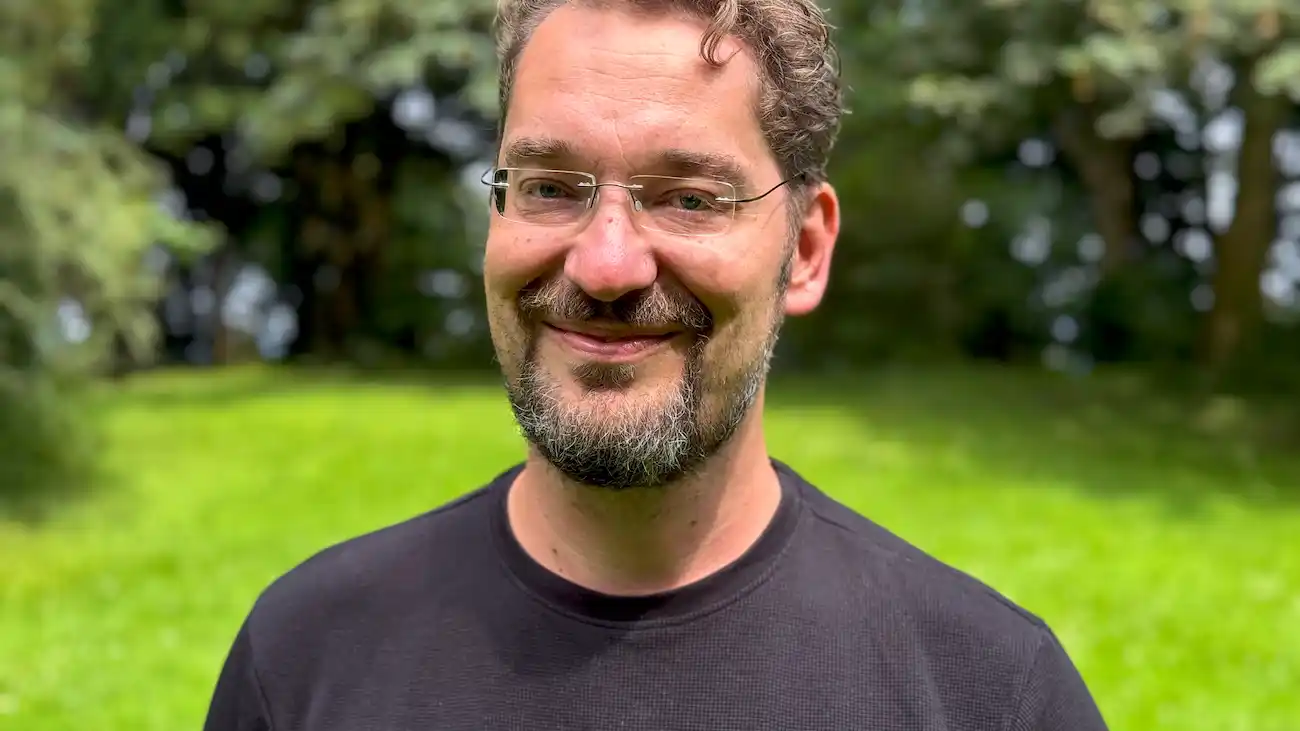 Porträt eines lächelnden Mannes mit Brille und schwarzem T-Shirt vor einem grünen, natürlichen Hintergrund. Das Bild zeigt Carlo Matic, einen erfahrenen Mehrfach-Unternehmer und Angel-Investor, der über Innovationen und Investitionen in Sport-Startups spricht.
