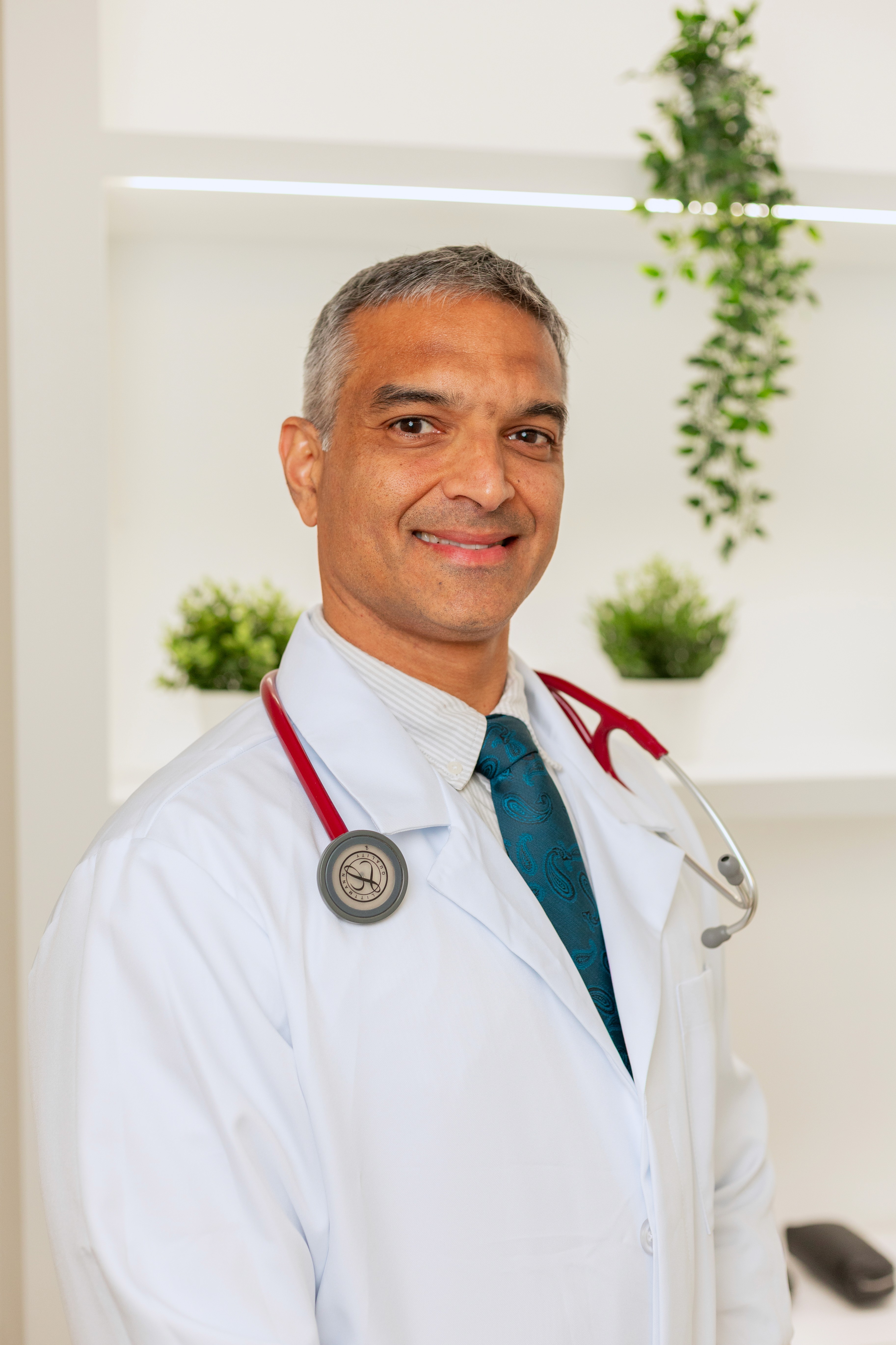 Smiling male doctor wearing a white coat and stethoscope, standing in a clinic setting with plants in the background.