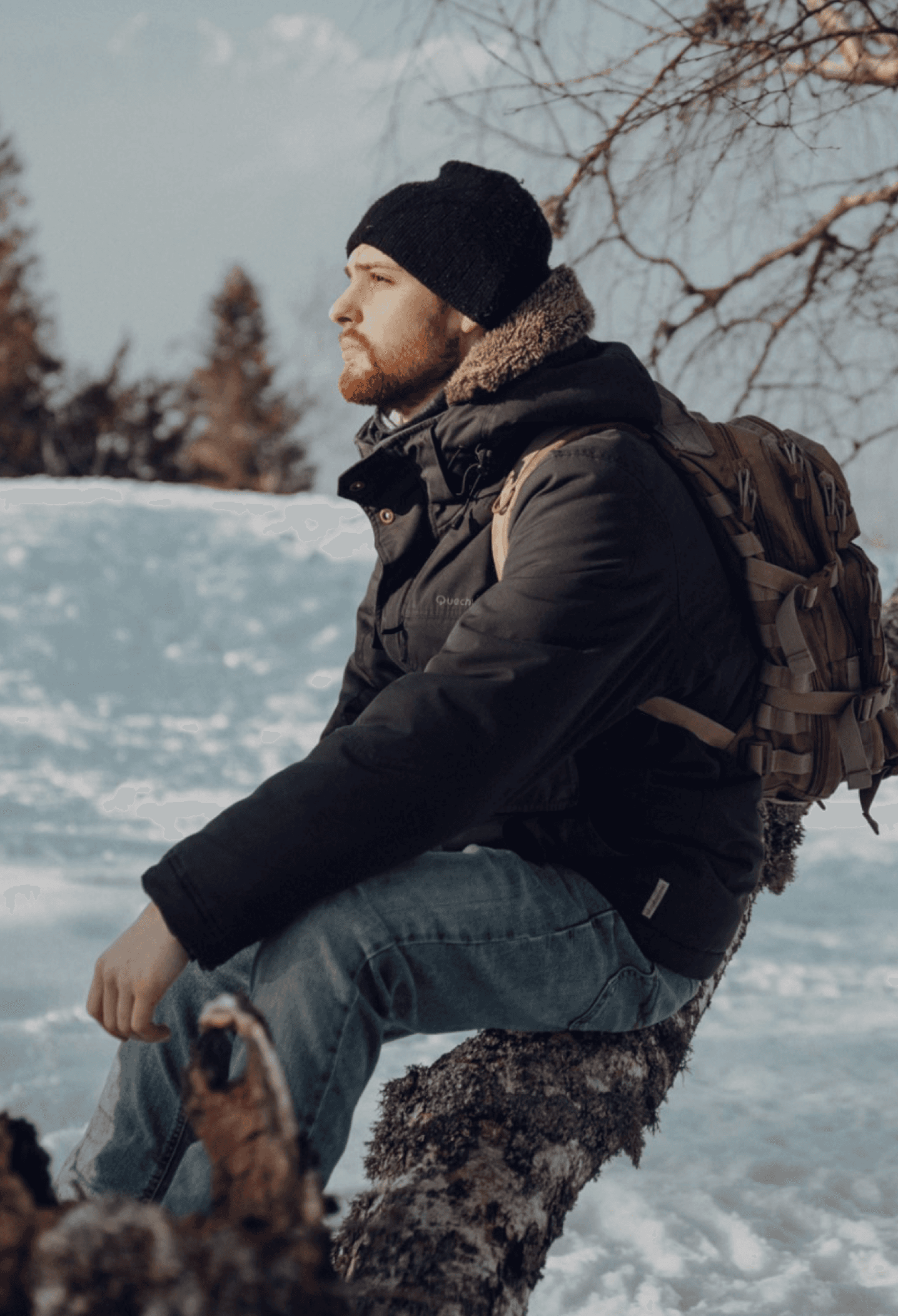 A man wearing a black jacket and a beanie sits on a tree branch in a snowy landscape, gazing thoughtfully into the distance. Bare tree branches are visible in the foreground.