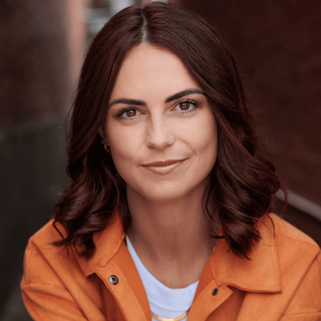 A woman with short curly hair wearing a black t-shirt looks into the camera.
