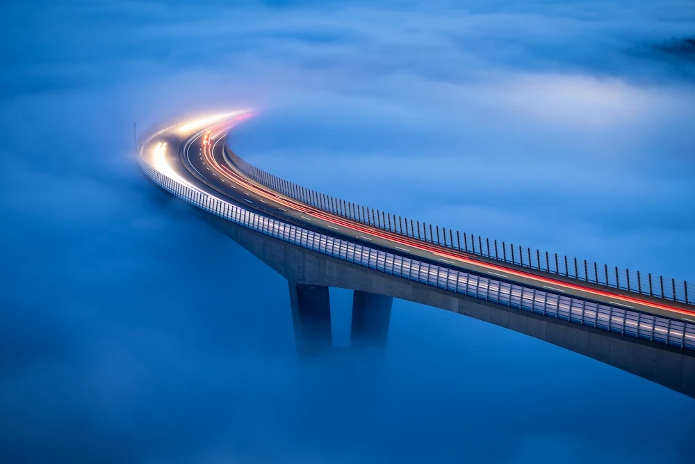 Aerial view of the Črni Kal viaduct in Slovenia at sunset, with its tall concrete pillars rising above a thick sea of fog in the valley during blue hour.