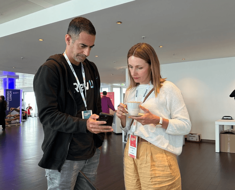 Two professionals networking at a tech conference; a man in a black hoodie showing his phone to a woman holding a coffee cup.