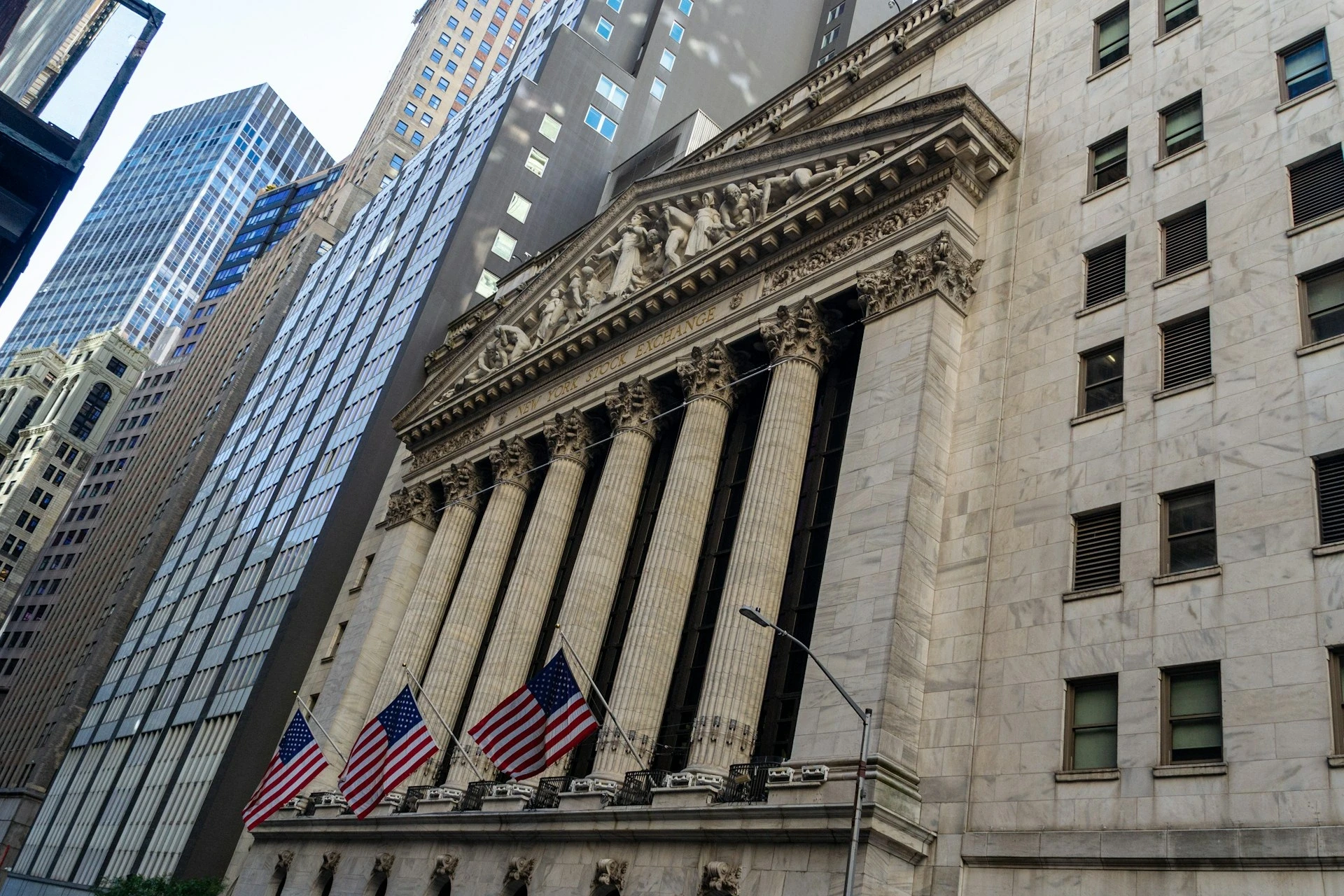 The building in the image is the New York Stock Exchange (NYSE) located on Wall Street in New York City. It’s recognizable by its grand neoclassical architecture featuring tall Corinthian columns, a pediment with sculpted figures, and multiple American flags displayed in front.