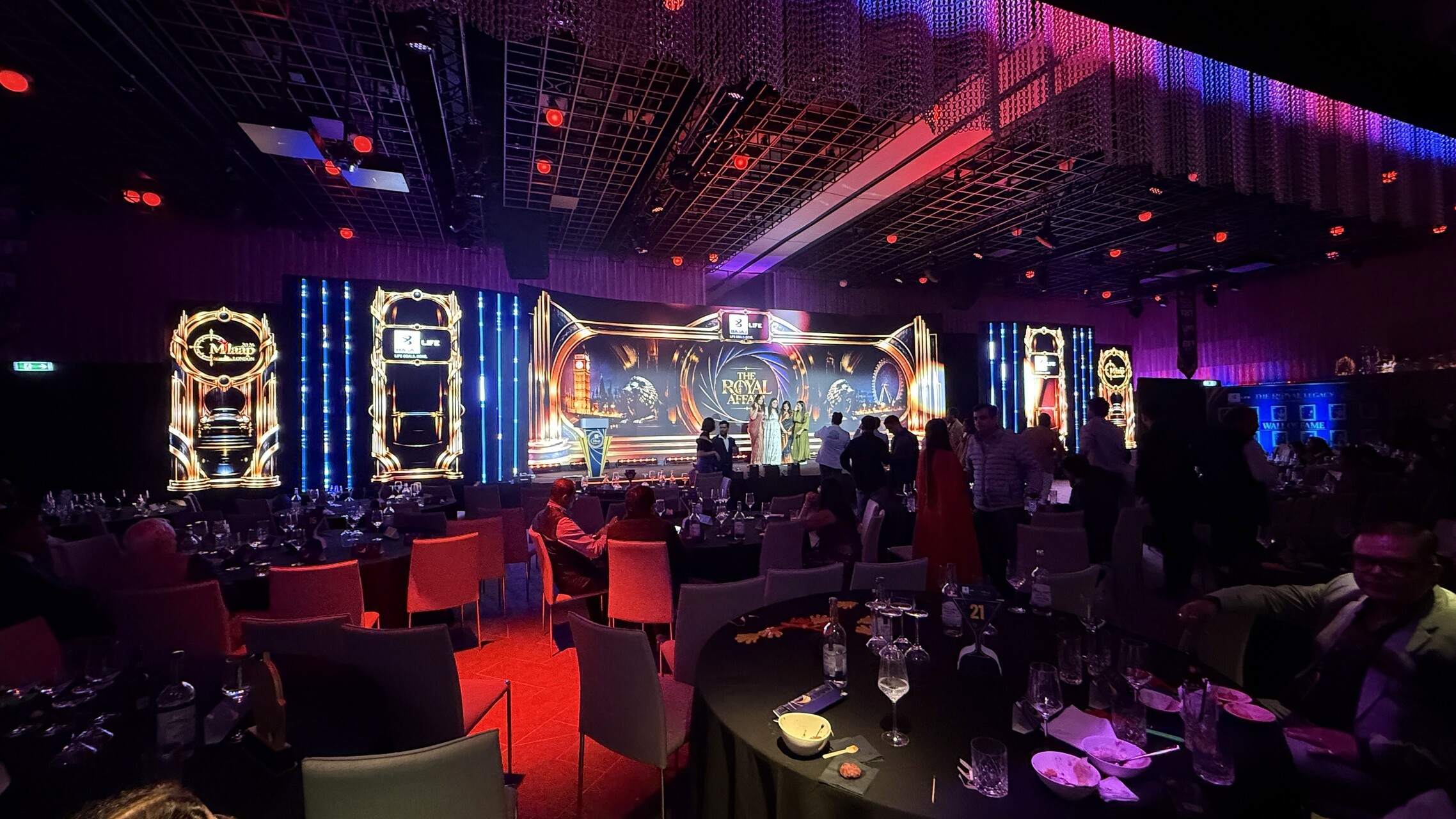 Wide view of the gala ballroom with gold stage visuals, blue FrameLED accents and guests beside dining tables.