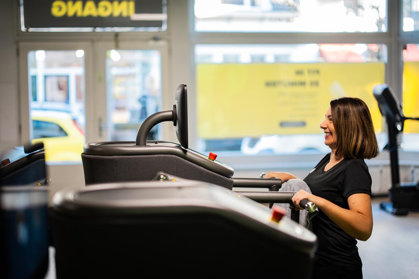 A woman with short hair stands at a treadmill in a gym, fitness equipment visible, bright windows in the background.