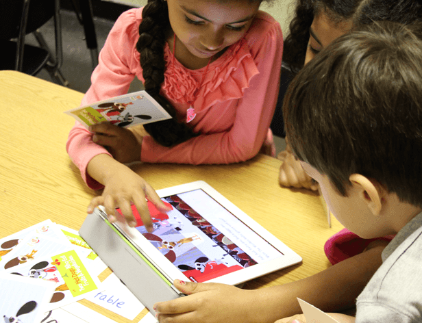 Three children are gathered around a light brown wooden table. The girl on the left, with long dark hair in a braid, wears a peach-colored long-sleeved shirt and holds a Mickey Mouse-themed booklet. The boy on the right, with short dark hair, wears a light gray shirt and points at a tablet displaying a colorful cartoon image. Behind them, two other children with dark hair lean in, observing the tablet screen.