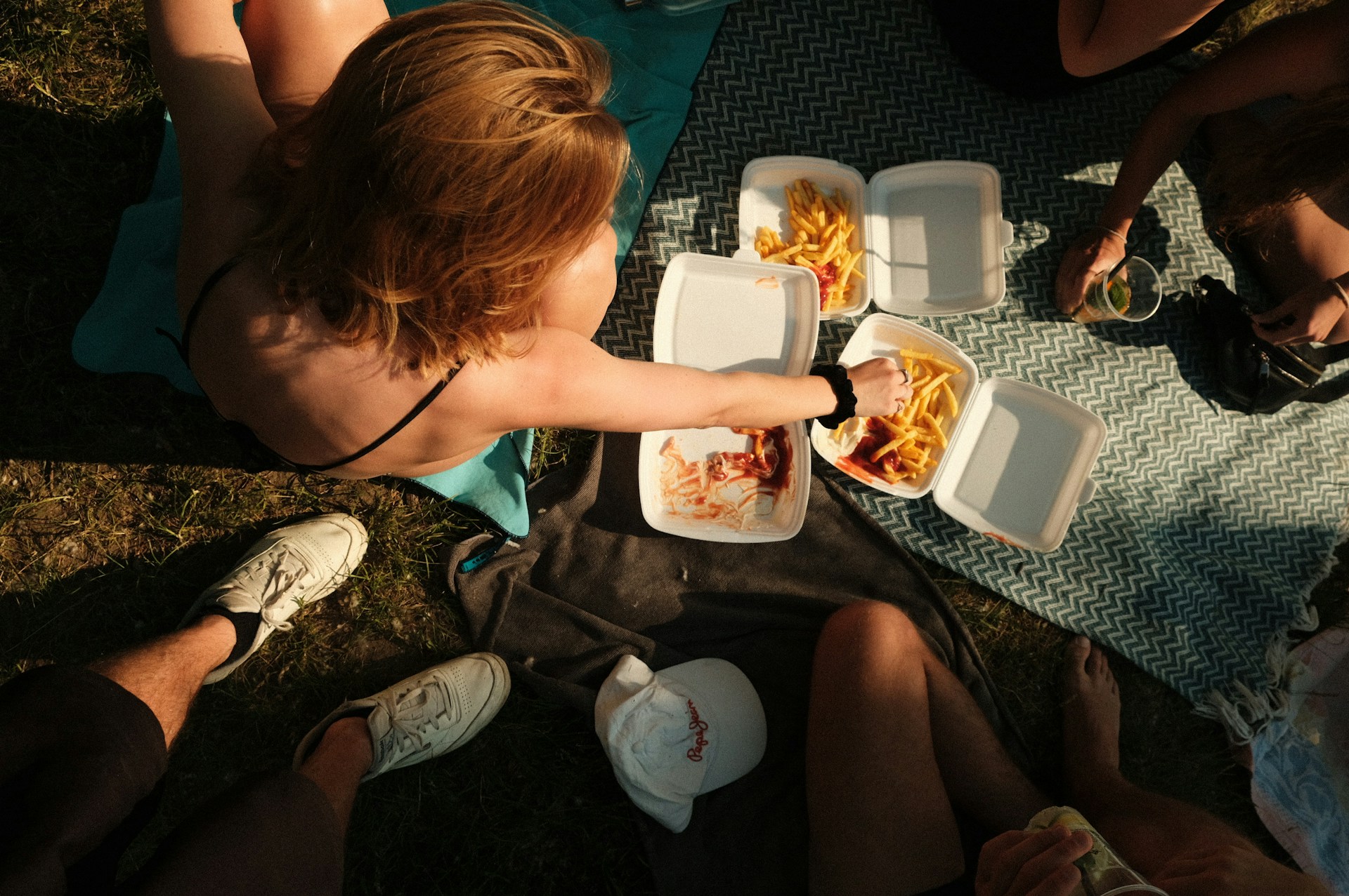 Top-down view of people having a sunny picnic on the grass, eating french fries with ketchup from white styrofoam takeout containers resting on blankets.