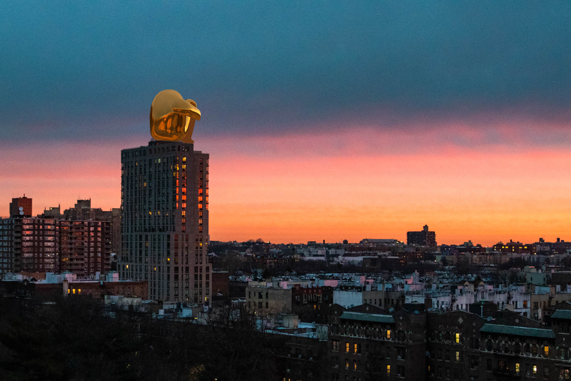 A tall, rectangular skyscraper with a gold-colored sculpture on top stands prominently against a vibrant sunset sky. The sky transitions from deep blue at the top to warm orange and pink hues near the horizon. The building’s facade features numerous windows, and its silhouette is visible against the colorful backdrop. In the foreground, a mix of residential buildings with varied roof colors and heights can be seen, extending into the distance.