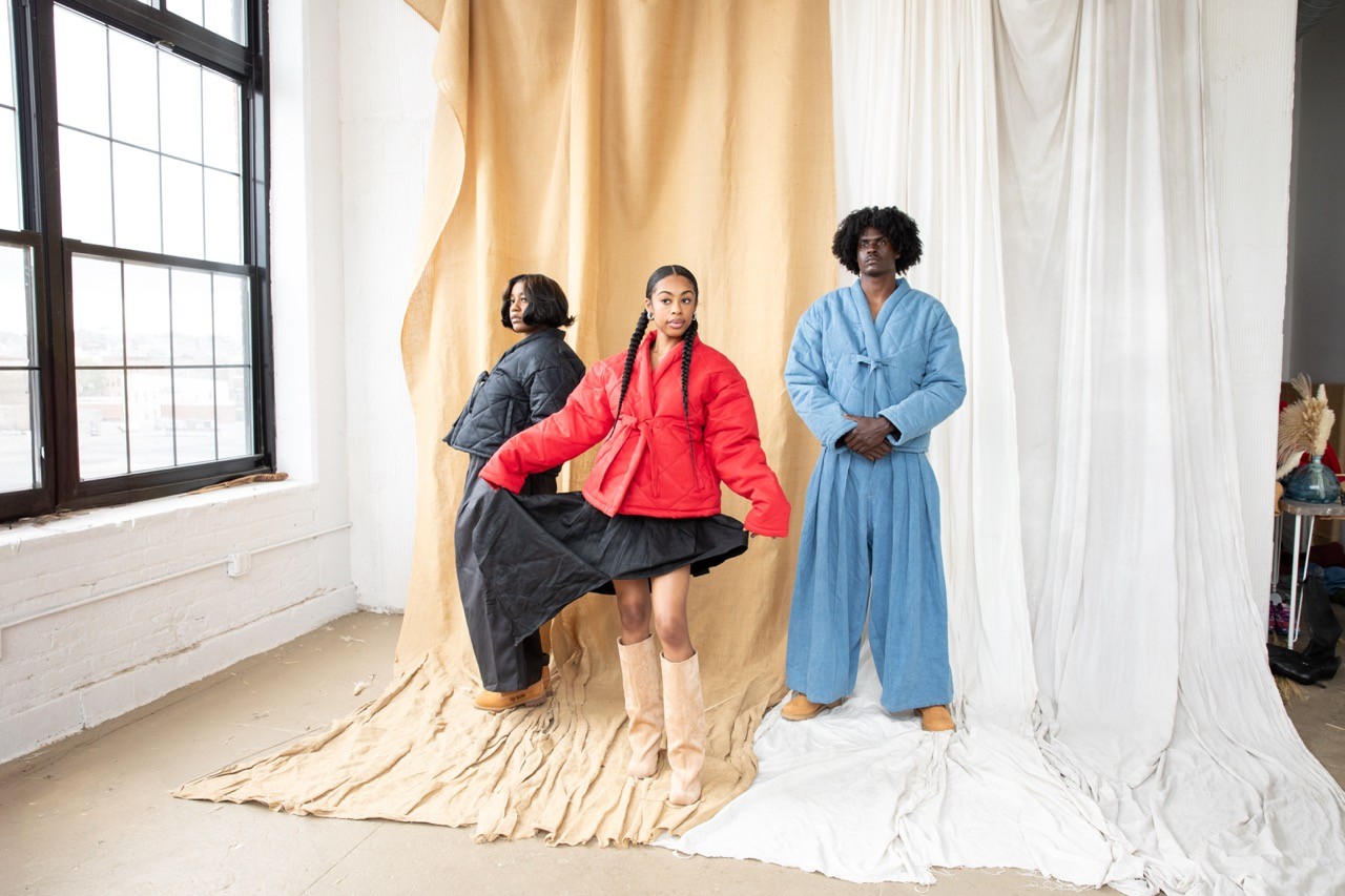 Three models posing in a studio with draped fabric backdrops, featuring a red jacket look centered in frame.