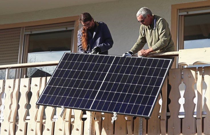 Zwei Personen installieren gemeinsam ein Solarpanel auf einem Balkon bei hellem Sonnenlicht.