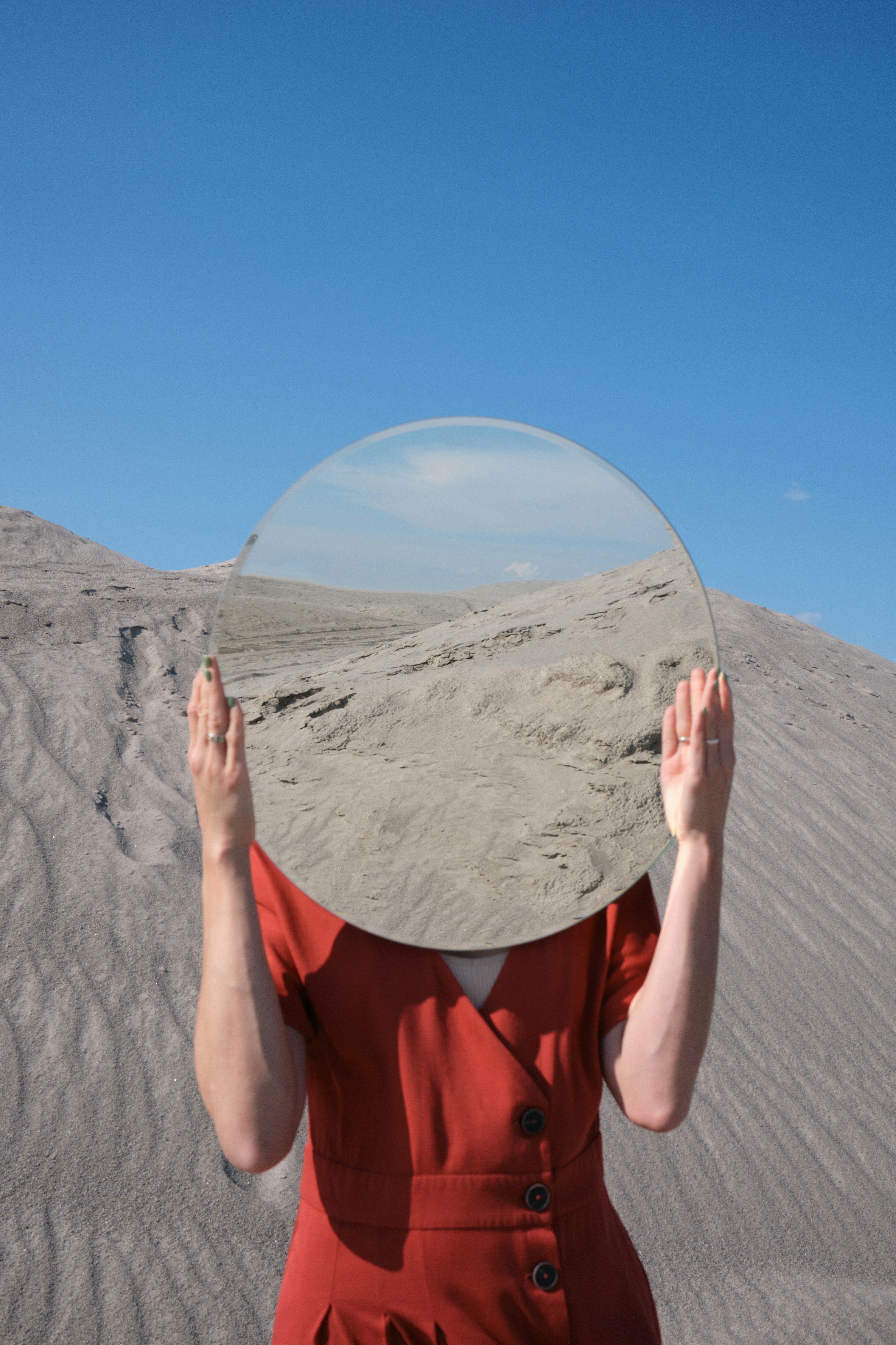 A person in a red dress holds a circular mirror against a desert landscape under a clear blue sky.
