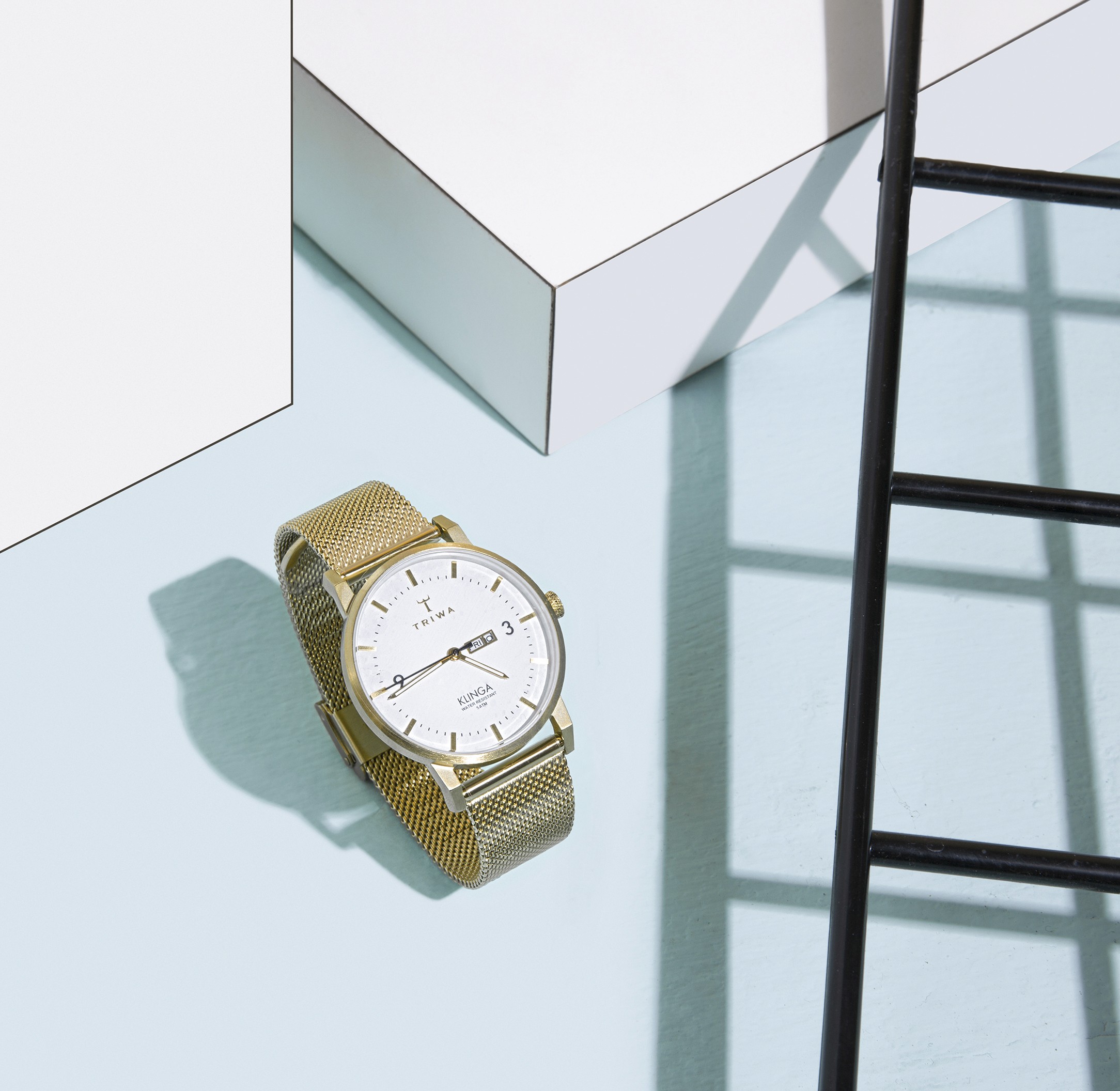 Men’s gold mesh strap watch from Florsheim Shoes displayed with minimalist geometric props casting shadows on a light blue surface.