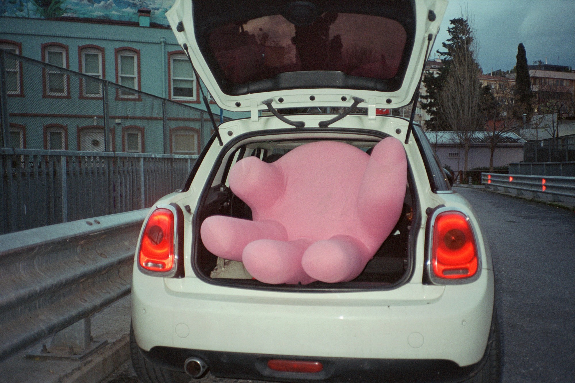 A large, pink plush figure is tightly packed in the open trunk of a white car parked beside a metal fence, with buildings and trees visible in the background.
