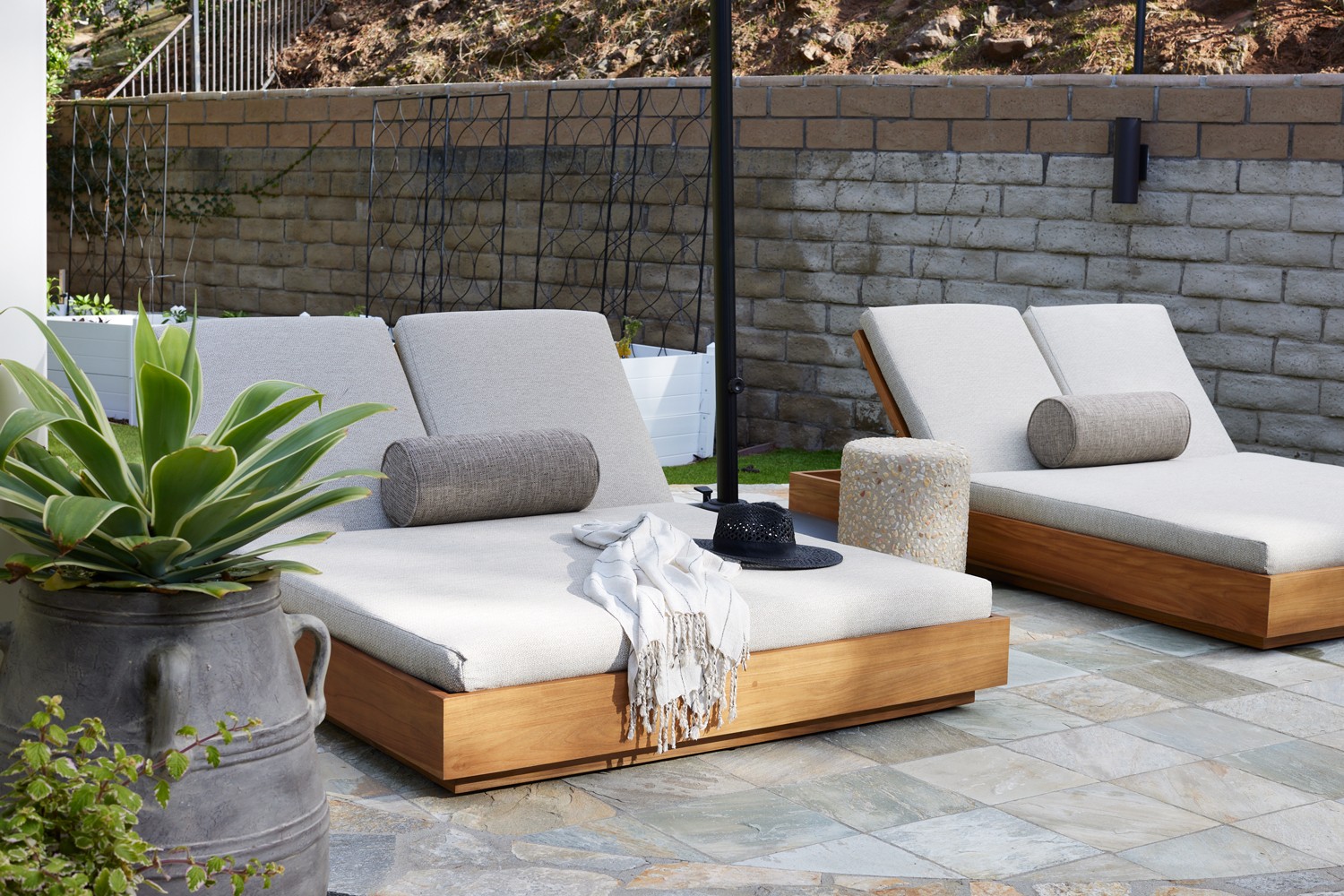 This outdoor oasis blends natural charm and refined luxury with a studded stone side table, linen accents, a sleek sun bed, large umbrella, and slate-inspired tiles, creating a harmonious sanctuary