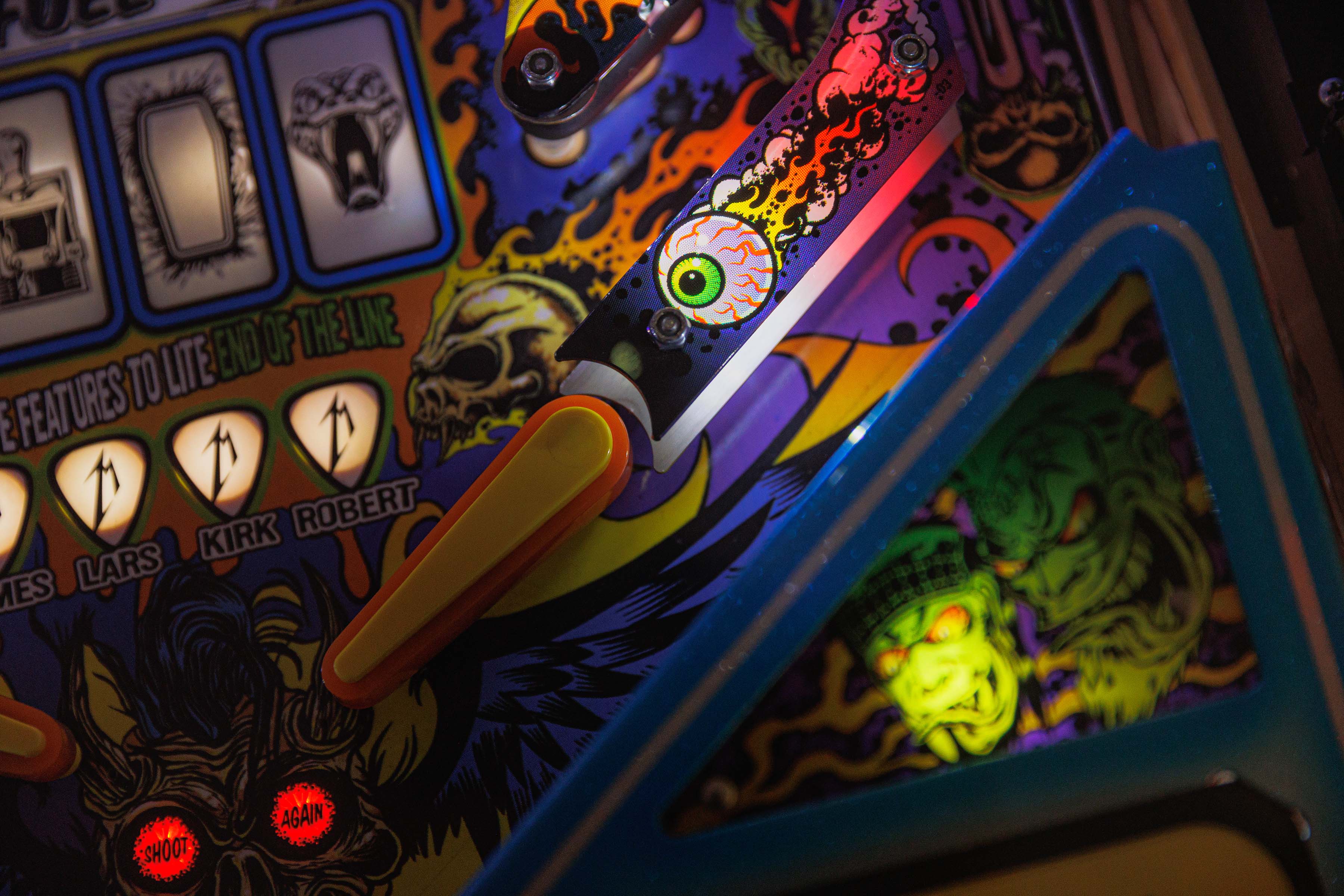 Close-up of a Metallica pinball machine’s flippers, featuring colorful monster art, flaming eyeball graphics, skull illustrations, and illuminated “Shoot Again” lights.