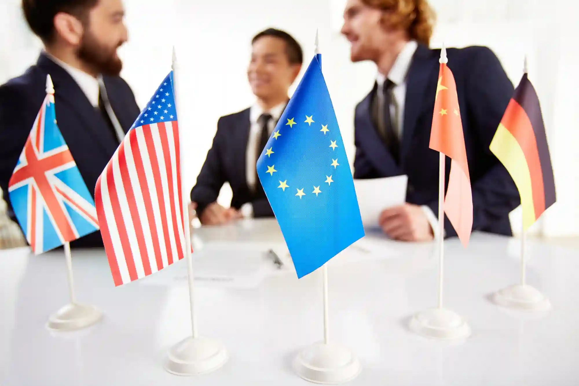 A row of international flags on a desk with businessmen blurred in the background, representing global digital policy negotiations.