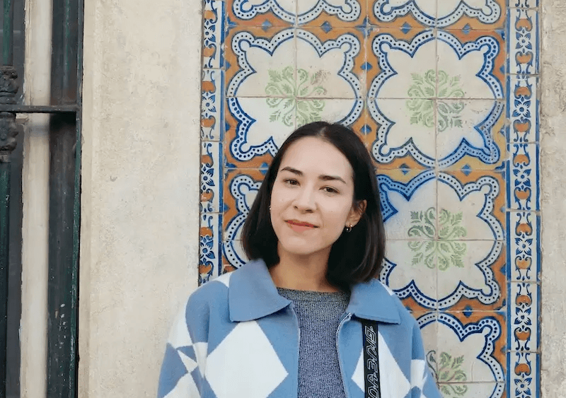 Caro smiling in a warm, outdoor setting with soft sunlight and a wall of painted Portuguese tiles in the background.