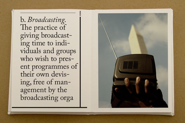 The image shows an open book with a definition of “broadcasting” on the left page and a photograph on the right. The definition reads: “The practice of giving broadcasting time to individuals and groups who wish to present programmes of their own devising, free of management by the broadcasting orga.” The photograph depicts a hand holding a black mobile phone with an antenna; the hand is positioned against a light-colored background that includes the Washington Monument. The phone is the central focus of the image.