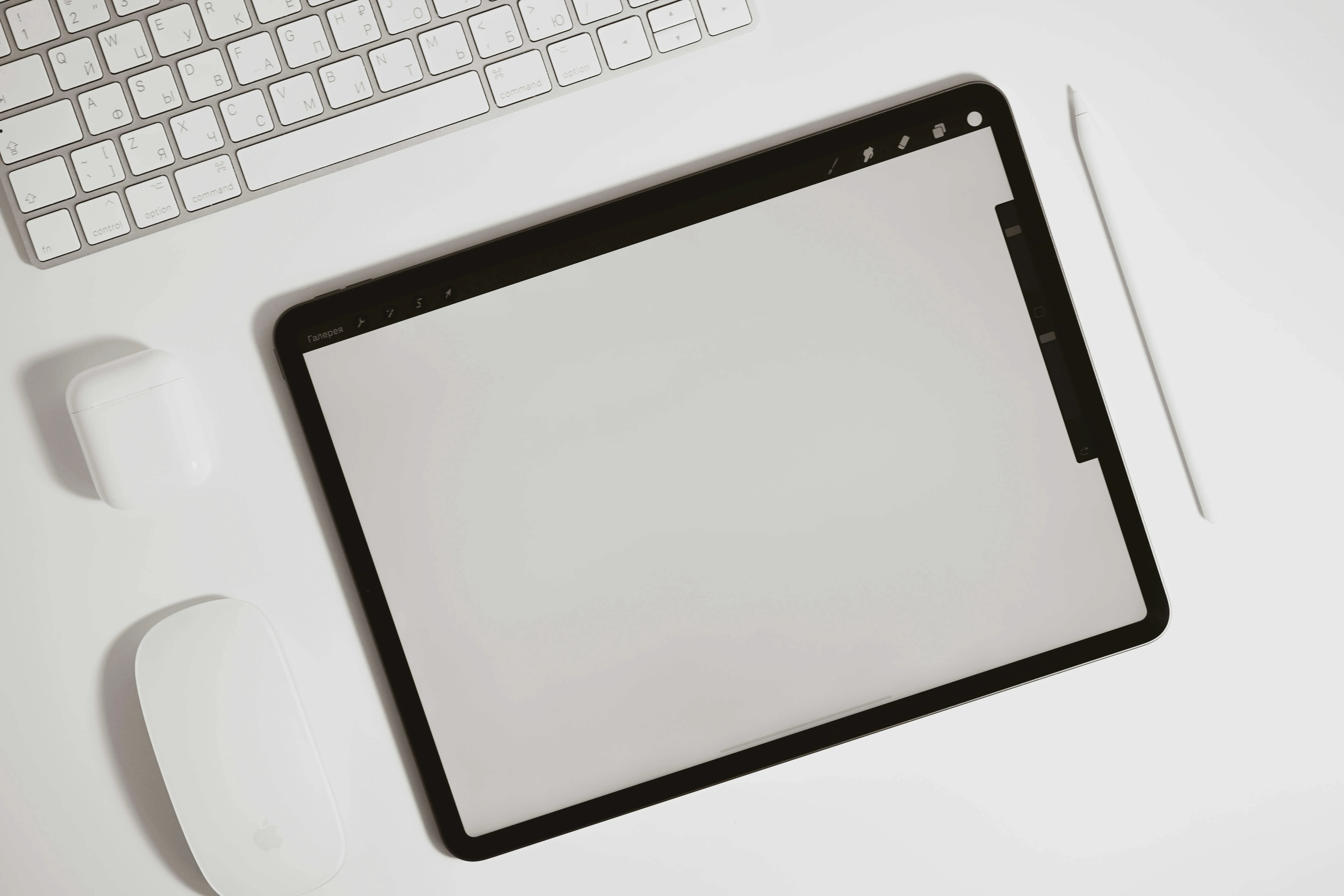 iPad with Apple Pencil next to a white Apple keyboard, AirPods, and Magic Mouse on a clean white desk.