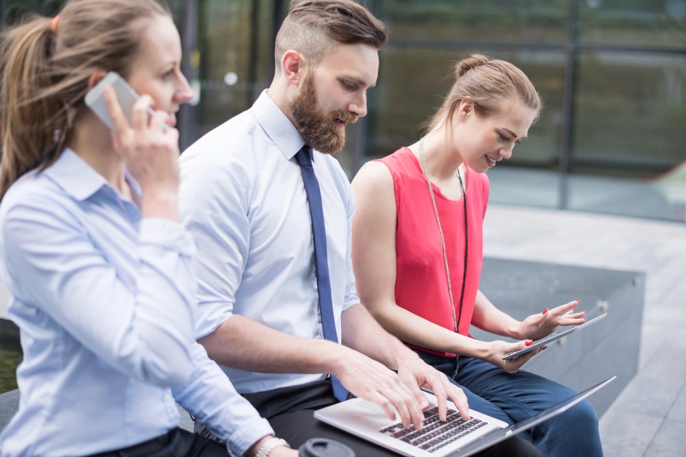 Three business professionals seated on a bench, each using a laptop, engaged in a collaborative work session outdoors.