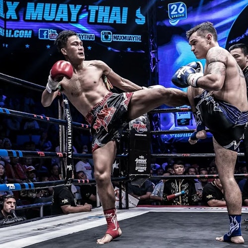 在擂台上有两名泰拳拳手，其中一名正在向另一名拳手的躯干发出高踢，背景中有观众和横幅。