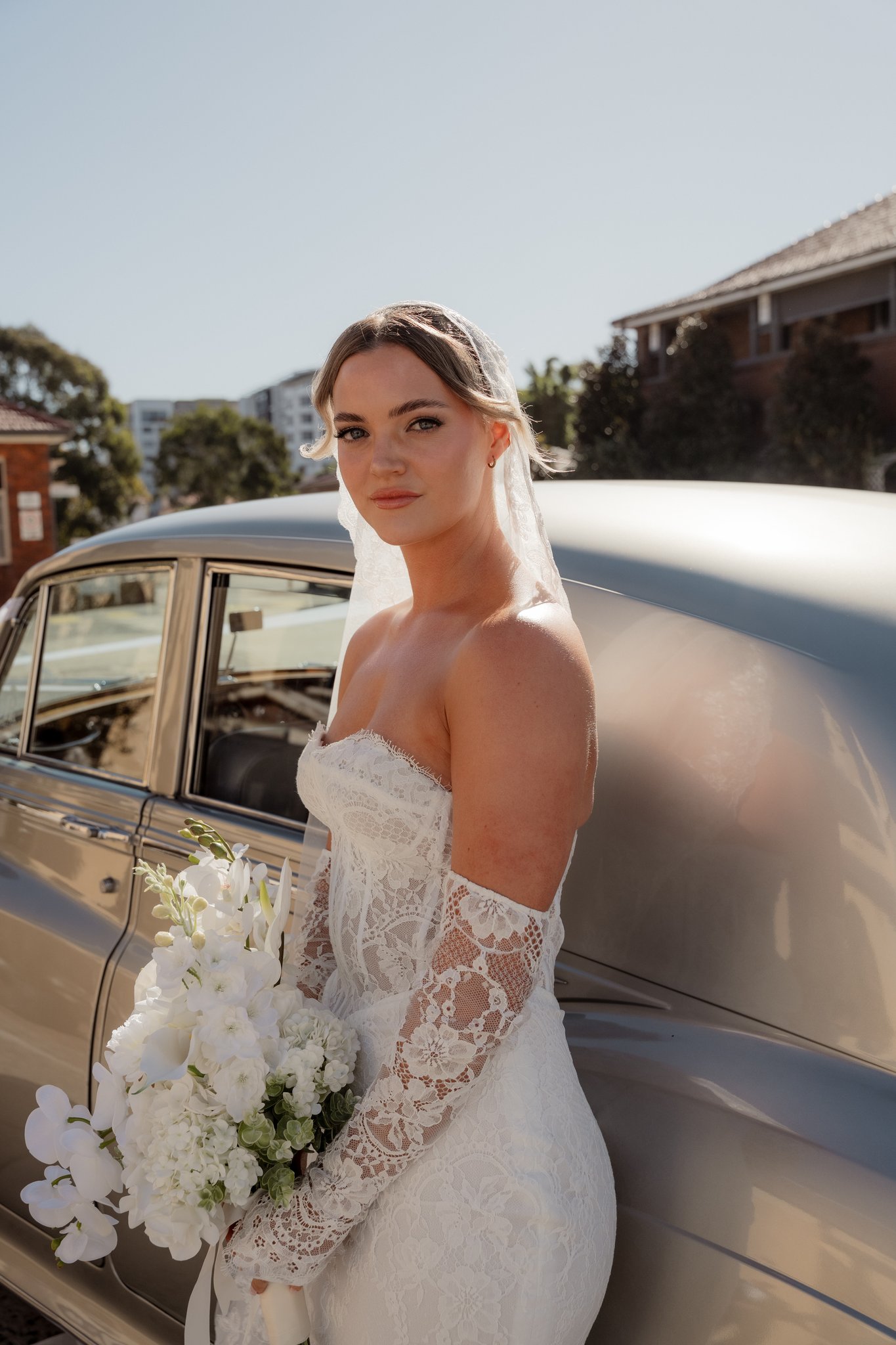 Bride in strapless lace gown and veil holds a white bouquet beside a vintage silver car.