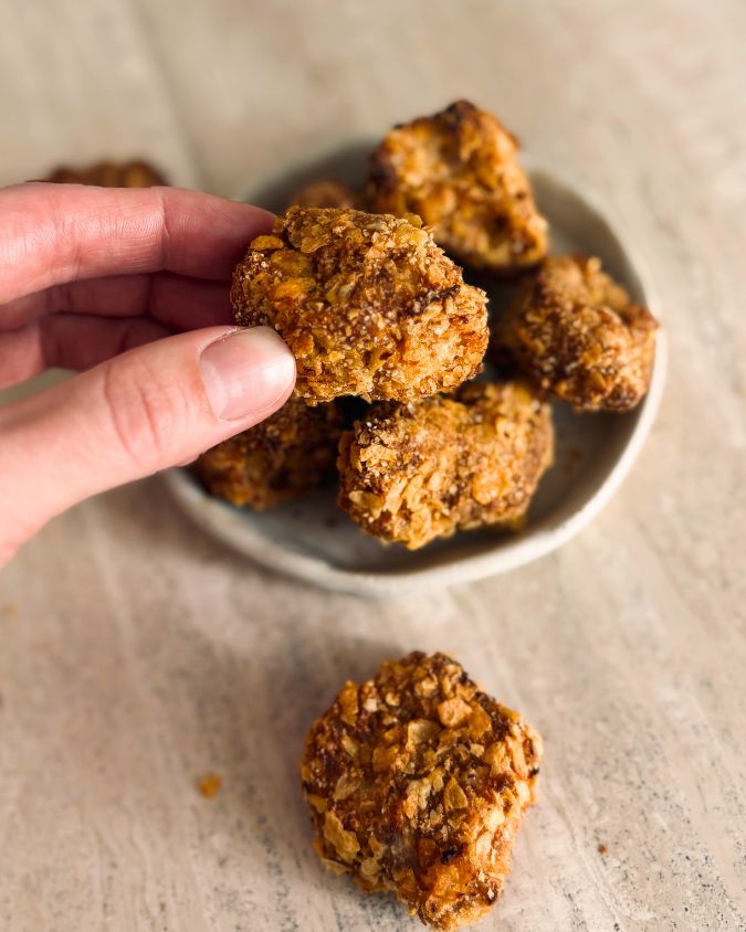 Hand holding a crispy tofu nugget over a small ceramic bowl filled with more nuggets on a wooden surface.