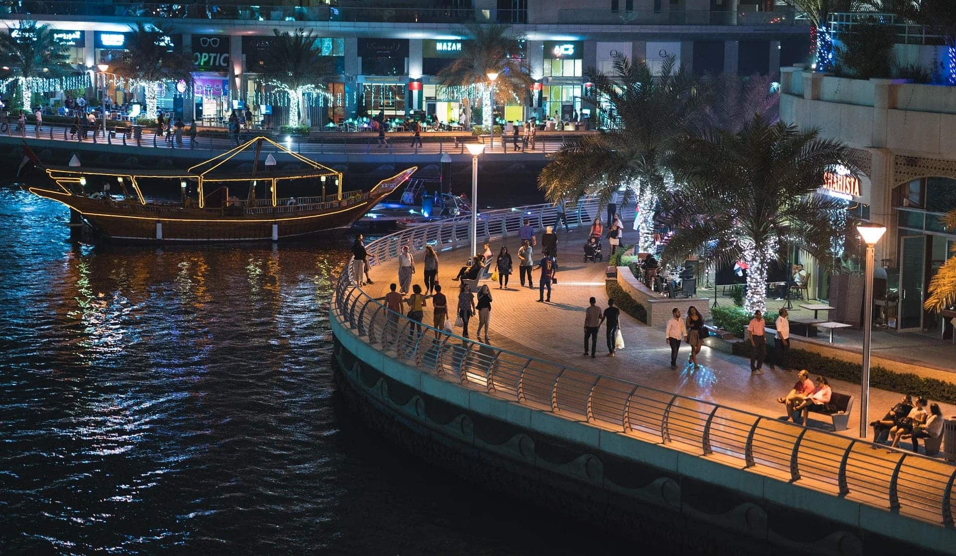 People walking along the Dubai Marina promenade at night, with waterfront restaurants, palm trees, and a lit dhow boat on the canal.
