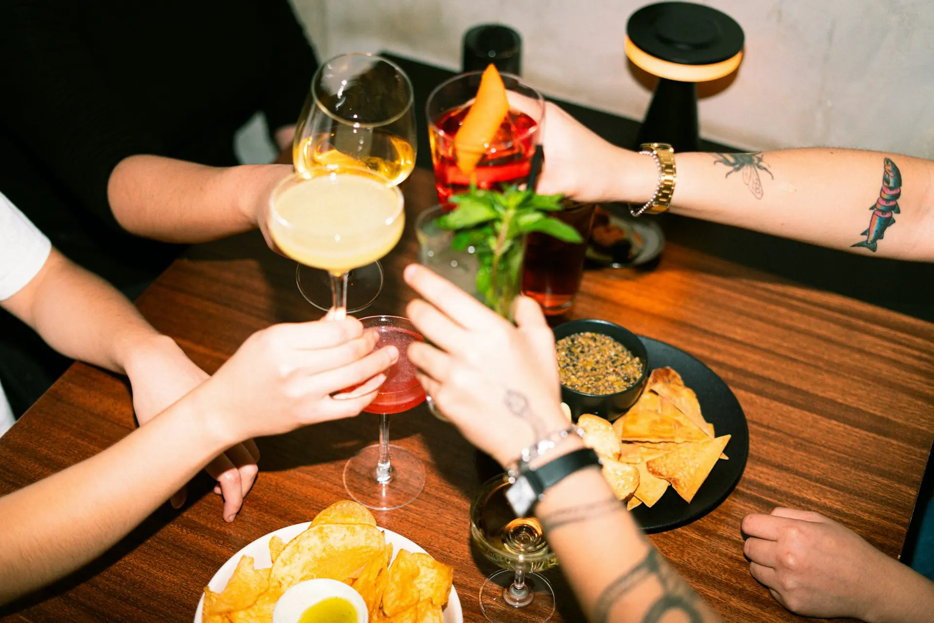 Friends clinking cocktail glasses over a wooden bar table with chips and dips, colorful drinks, and warm ambient lighting in a casual bar setting.