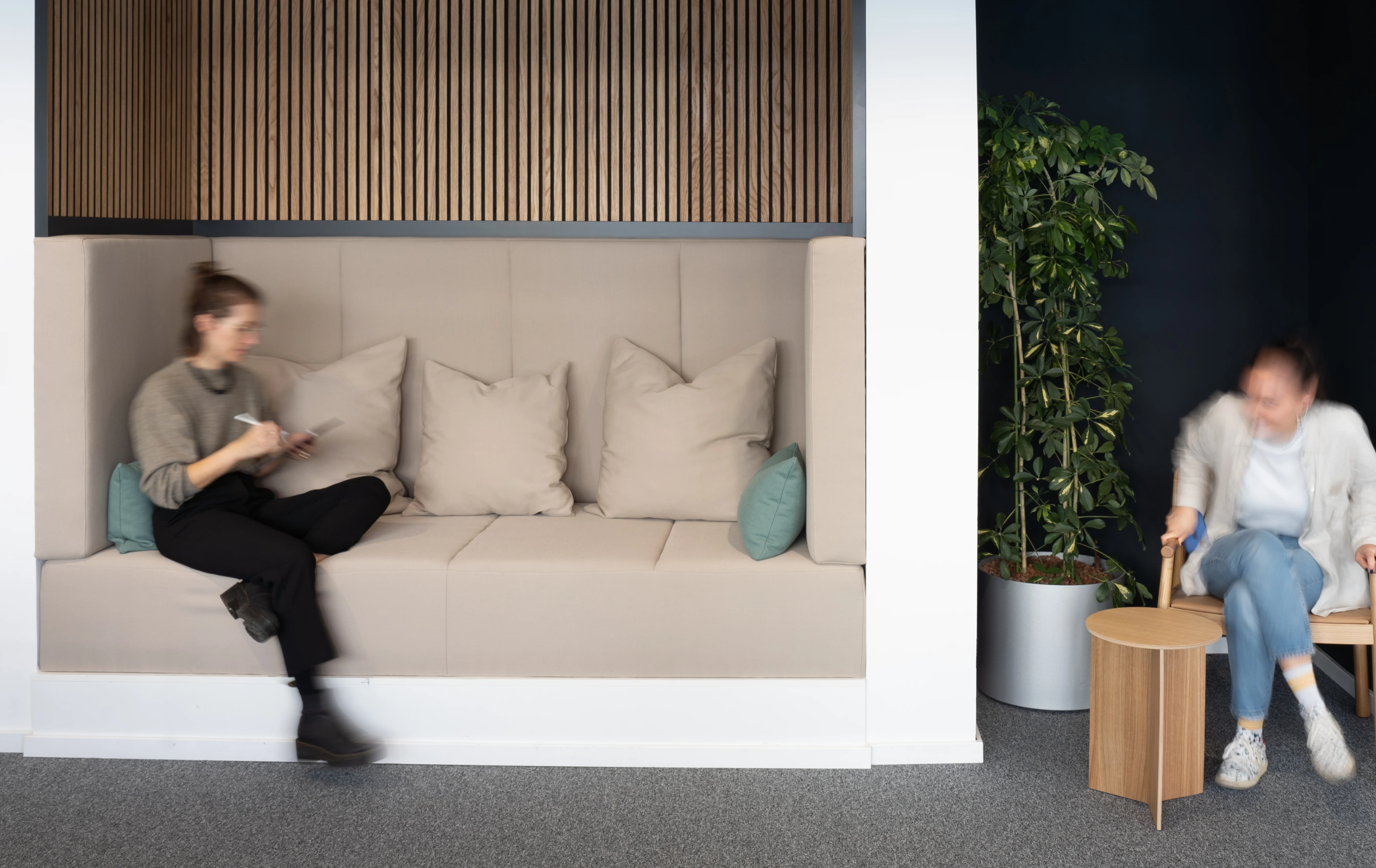 High-backed alcove sofa at AI START-UP MUNICH in beige with mint cushions under an oak slat canopy, one person reading on the bench and another seated by a planted corner.