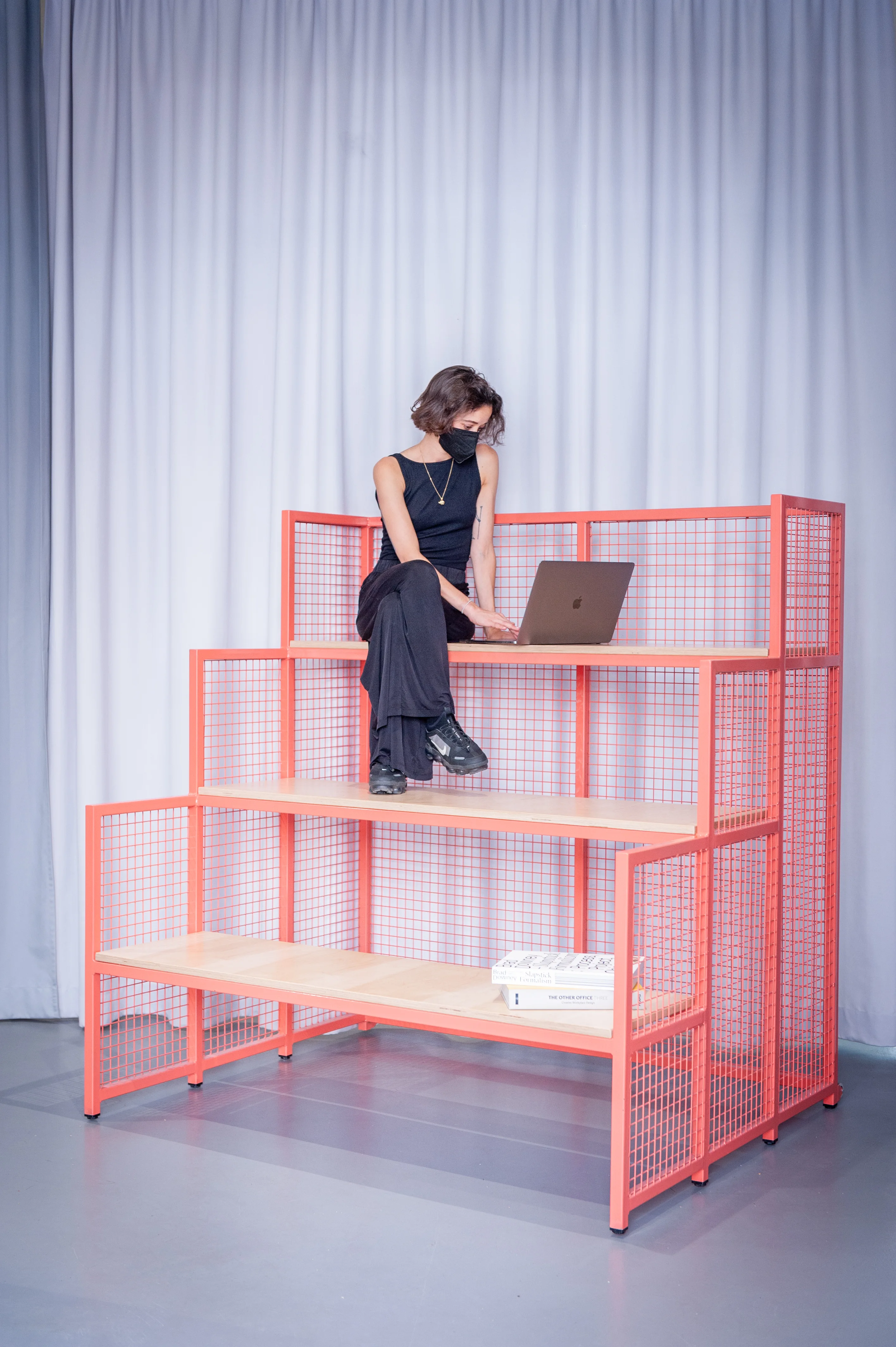 Woman sitting with a laptop on the top step of a coral wire-mesh tribune module at HEY CONTENT STUDIO in front of grey curtains.