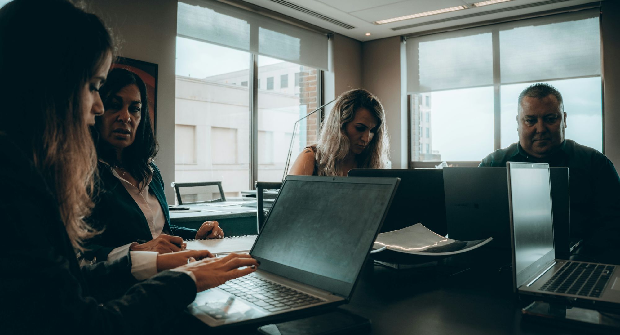imagem em tom escuro de pessoas sentadas ao redor de uma mesa trabalhando em notebooks