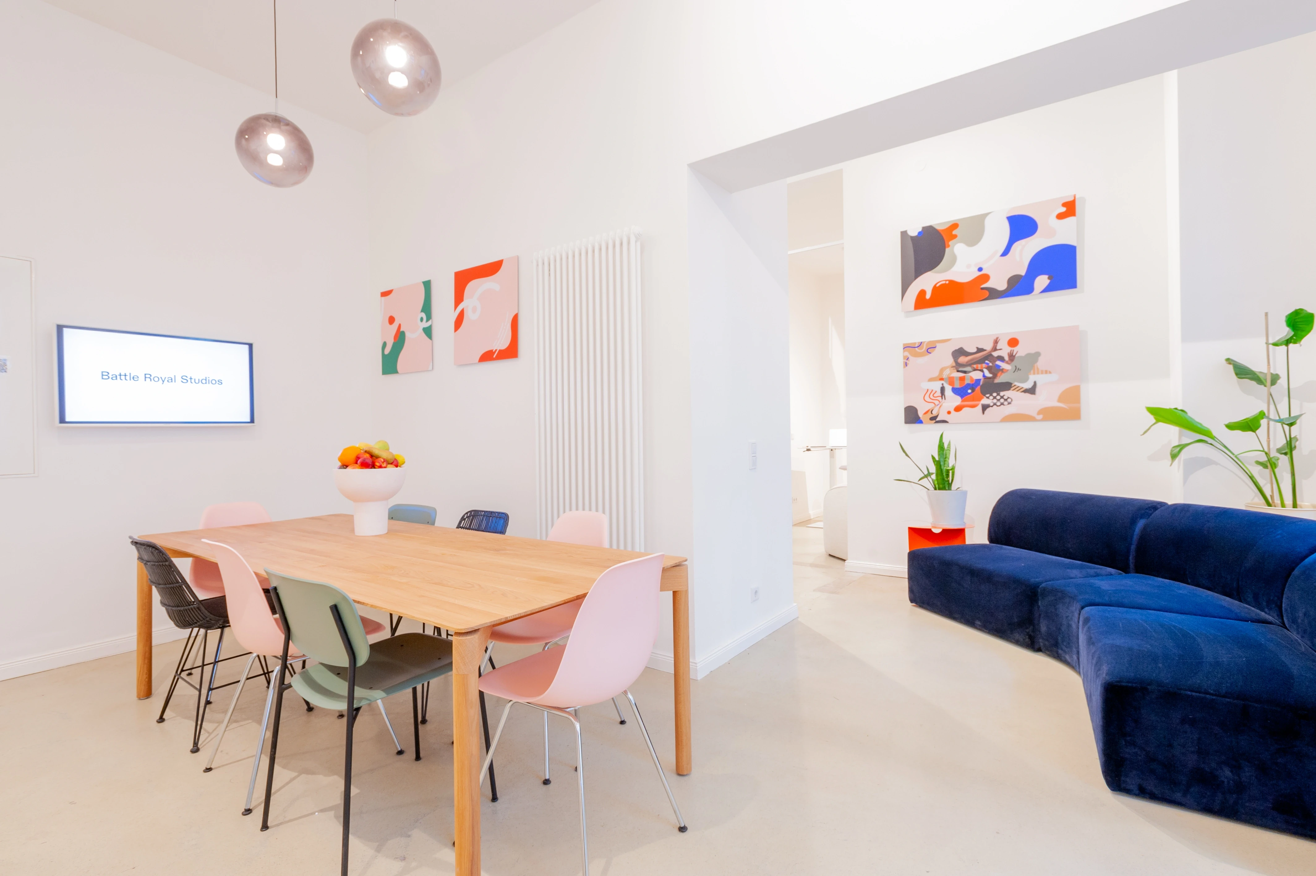 Lounge and dining area at BATTLE ROYAL with a wooden table and pastel chairs in pink, green and navy, a fruit bowl, abstract wall art, glass pendant lights and a curved navy velvet sofa.