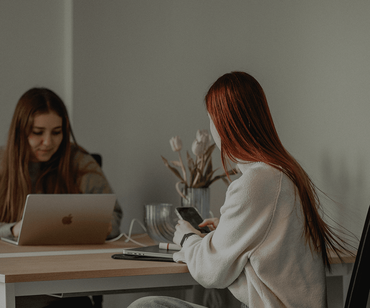 Zwei Personen sitzen an einem hellen Schreibtisch in einem Coworking Space. Die Person im Vordergrund trägt einen cremefarbenen Pullover und weite Jeans, hat lange rotbraune Haare und hält ein Smartphone in der Hand. Auf dem Tisch stehen ein Laptop, ein Glas Wasser und eine Vase mit hellen Tulpen. Die Umgebung wirkt modern und minimalistisch, mit neutralen Wandfarben und einem dunklen Boden.