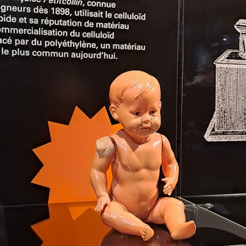 En sliten, naken plastdocka med en skadad axel visas mot en svart bakgrund med fransk text och illustrationer.