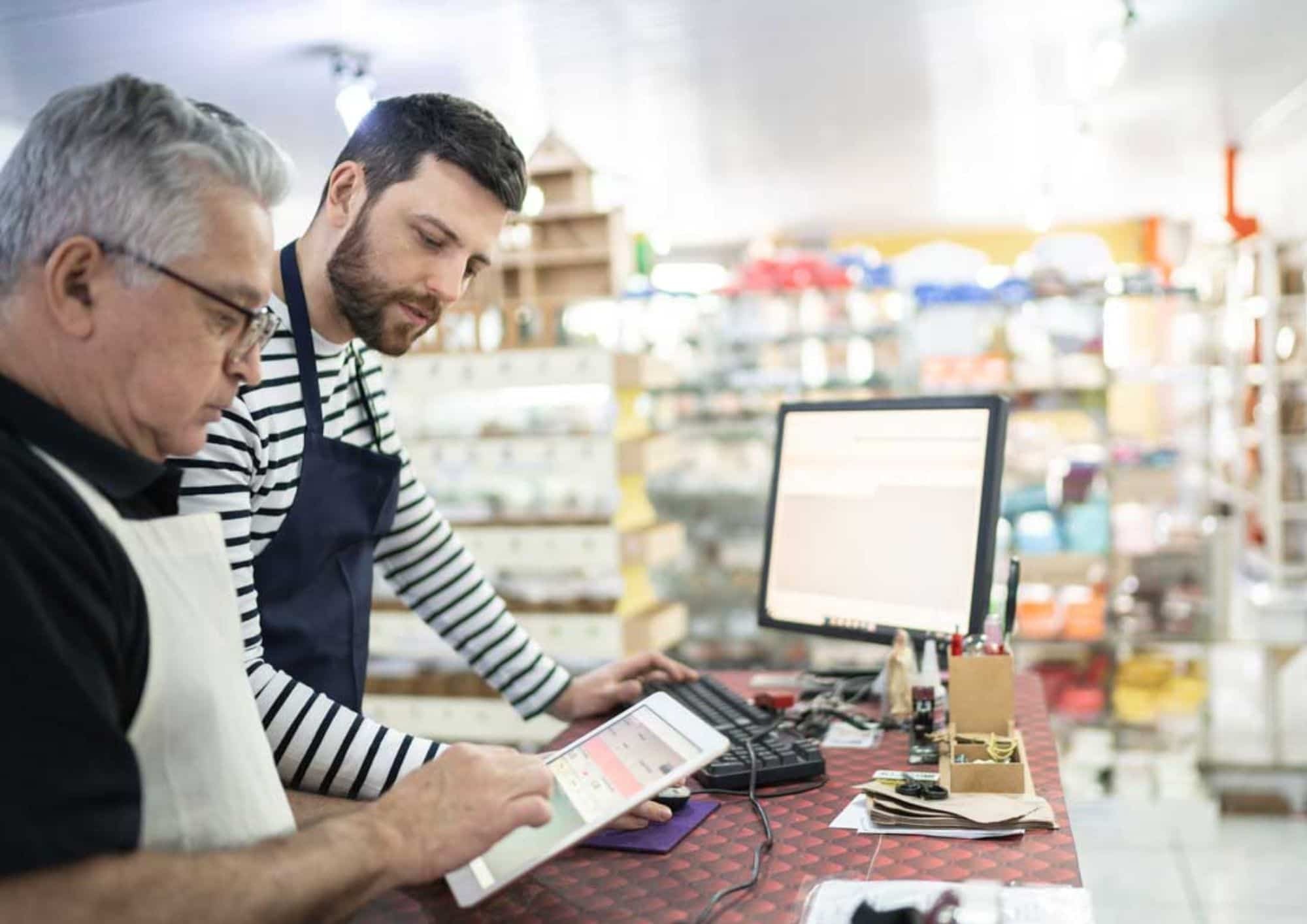 Duas pessoas no balcão de uma loja consultam informações em um tablet e em um computador. O ambiente comercial aparece ao fundo com prateleiras desfocadas.