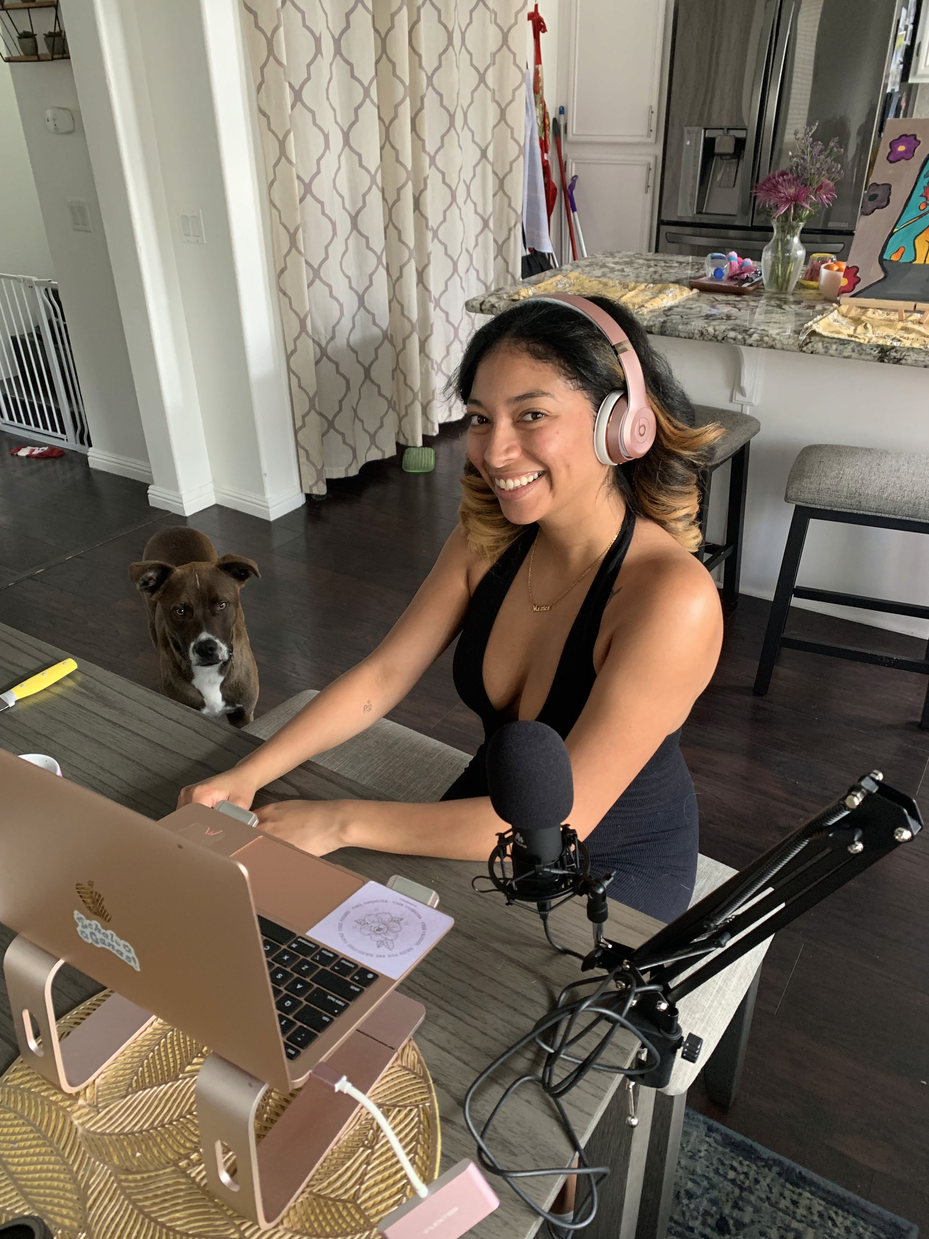 woman with pink headphones smiling nexct to a laptop and podcast mic and brown dog
