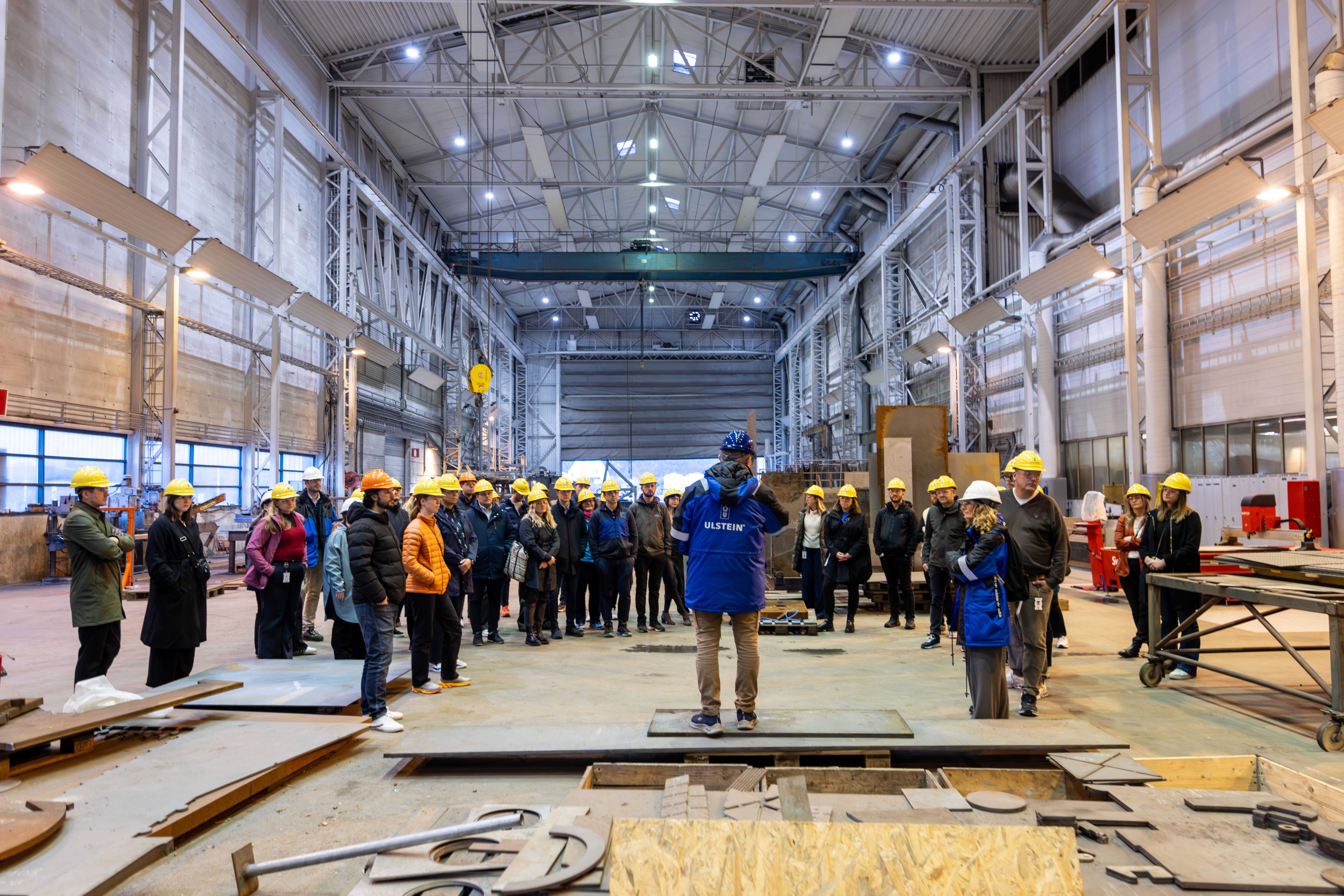 En gruppe mennesker i sikkerhetsvester inspiserer hyller i et stort lager. Atmosfæren er travel og profesjonell.